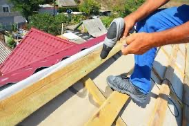 Pracownik w niebieskich spodniach przykręca drewnianą łatę do konstrukcji dachu pokrytego czerwoną blachodachówką za pomocą wkrętarki akumulatorowej, widok z góry.