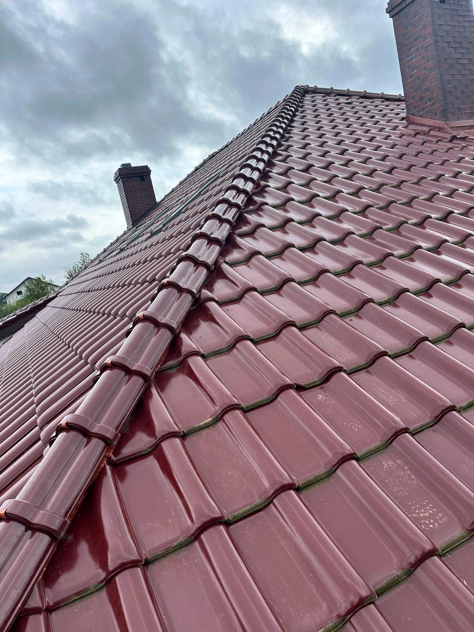 Błyszczący, bordowy dach z dachówki ceramicznej, widok z dołu na tle pochmurnego nieba, z kominem w tle.