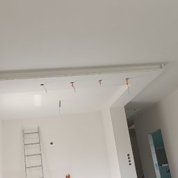 Krystian Leszczyński - Wnętrze w trakcie remontu: białe ściany, sufit z wnęką na oświetlenie, widoczne przewody elektryczne, drabina oparta o ścianę, okno zabezpieczone folią.