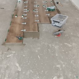 Krystian Leszczyński - Układanie podłogi z paneli imitujących drewno w korytarzu, widoczne narzędzia glazurnicze i poziomica laserowa.