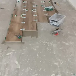 Układanie podłogi z paneli imitujących drewno w korytarzu, widoczne narzędzia glazurnicze i poziomica laserowa.