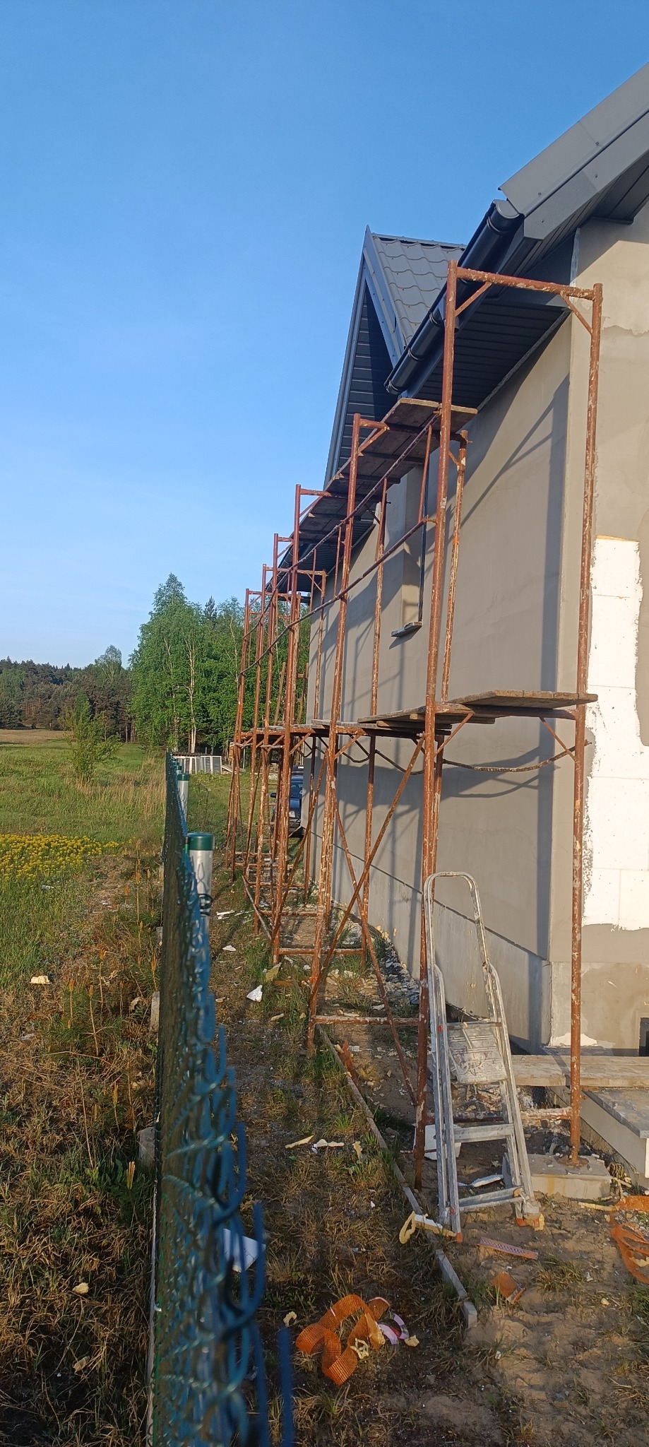 Ściana domu w trakcie prac wykończeniowych z rusztowaniem i drabiną, widoczne świeże tynki i częściowo ocieplenie, w tle zielona łąka i las.