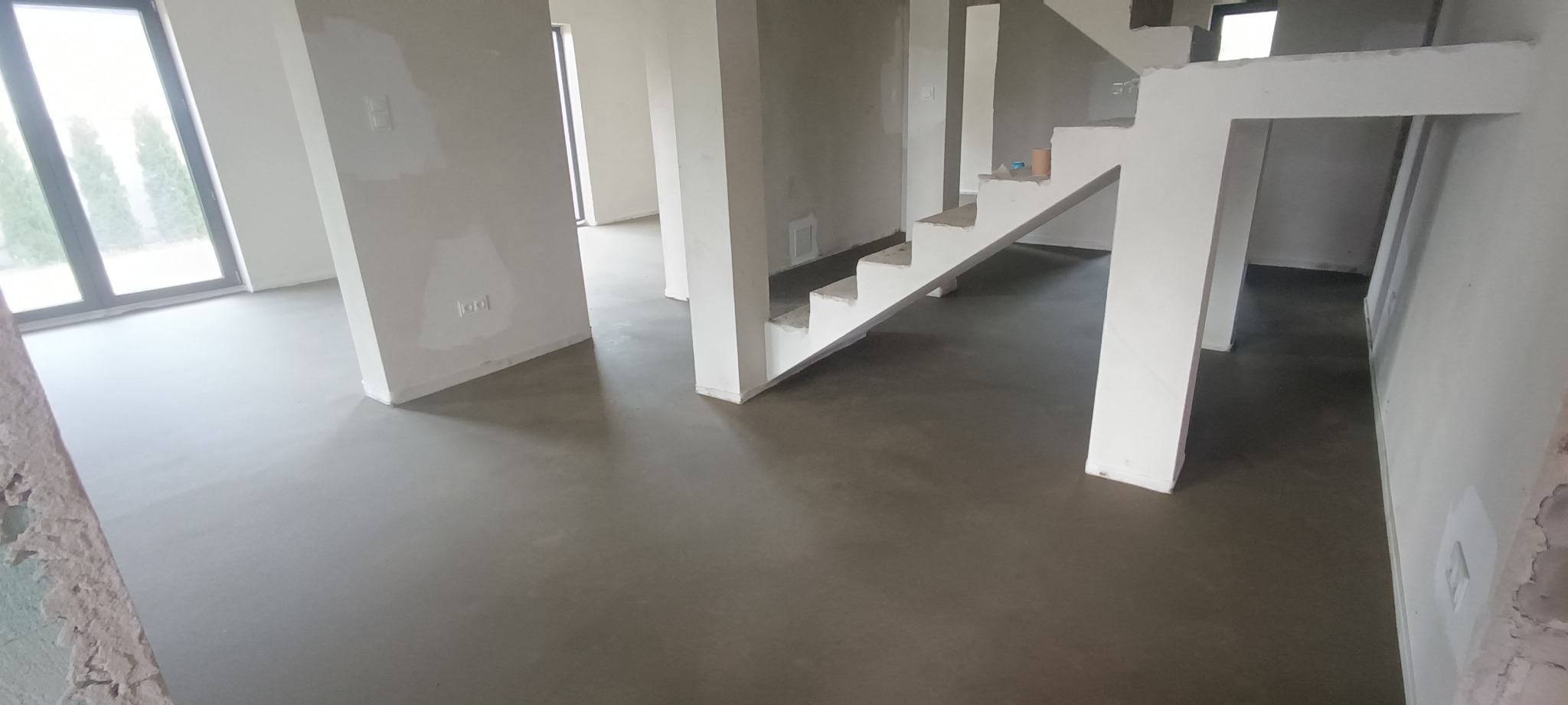 Surowy stan wnętrza domu z wylewką na podłodze, ścianami przygotowanymi do malowania i surowymi schodami bez balustrady.