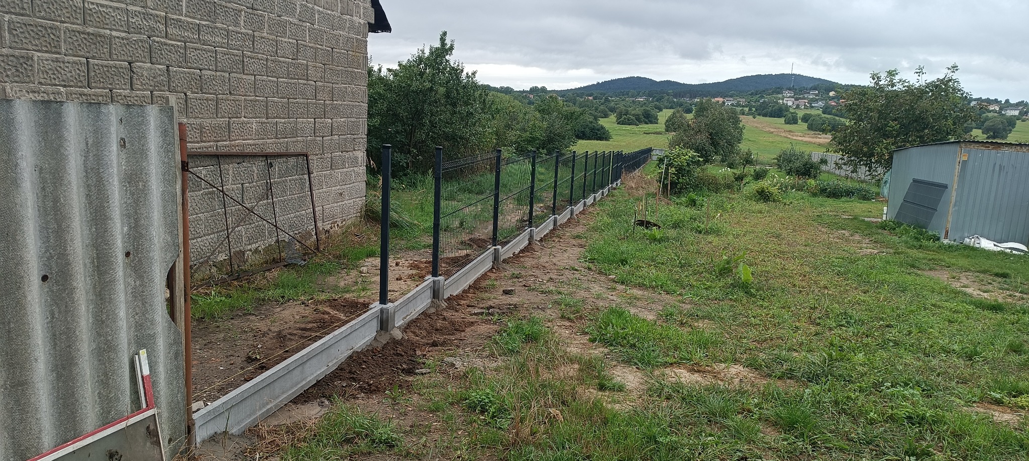 Montaż ogrodzenia panelowego z podmurówką betonową przy domu z elewacją z bloczków, widok na krajobraz zalesionych wzgórz w tle.