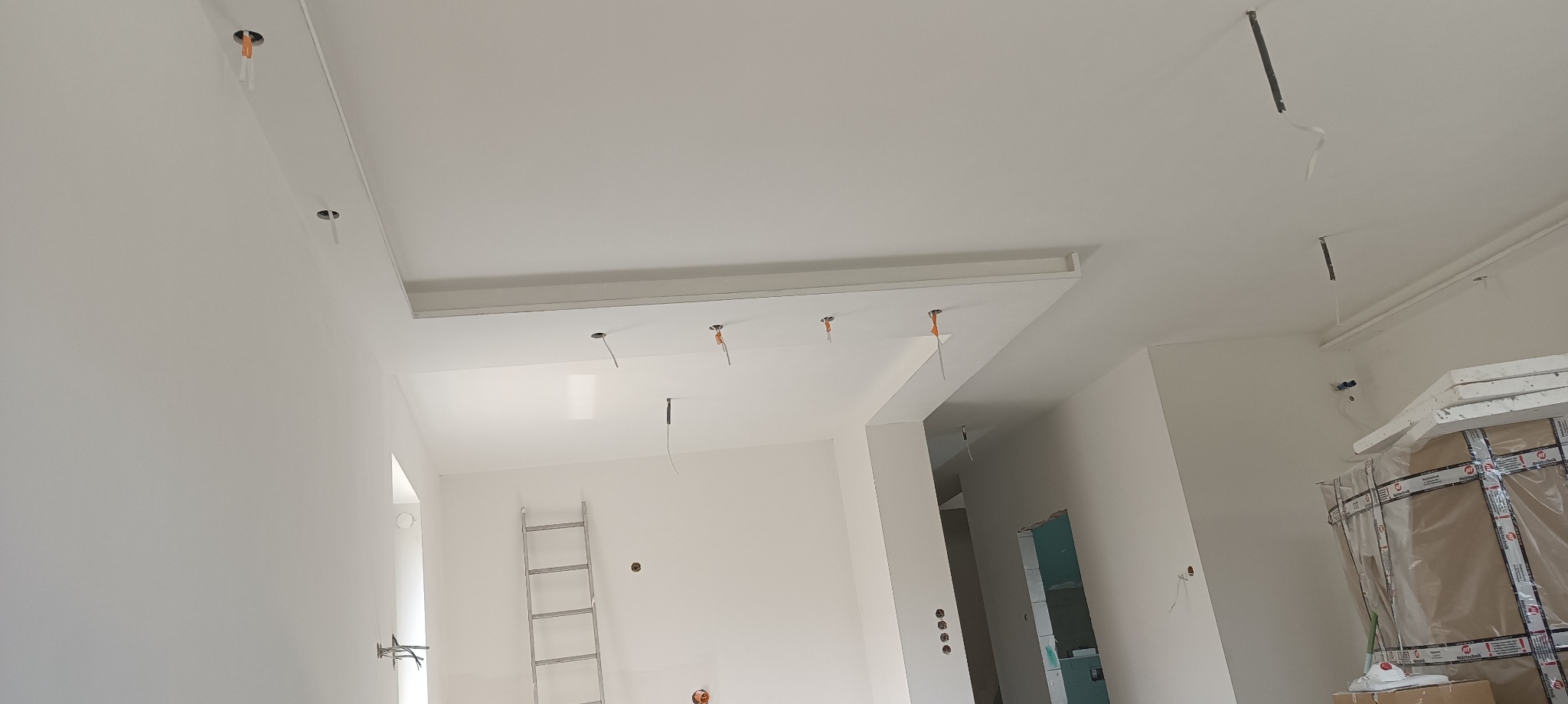 Wnętrze w trakcie remontu: białe ściany, sufit z wnęką na oświetlenie, widoczne przewody elektryczne, drabina oparta o ścianę, okno zabezpieczone folią.