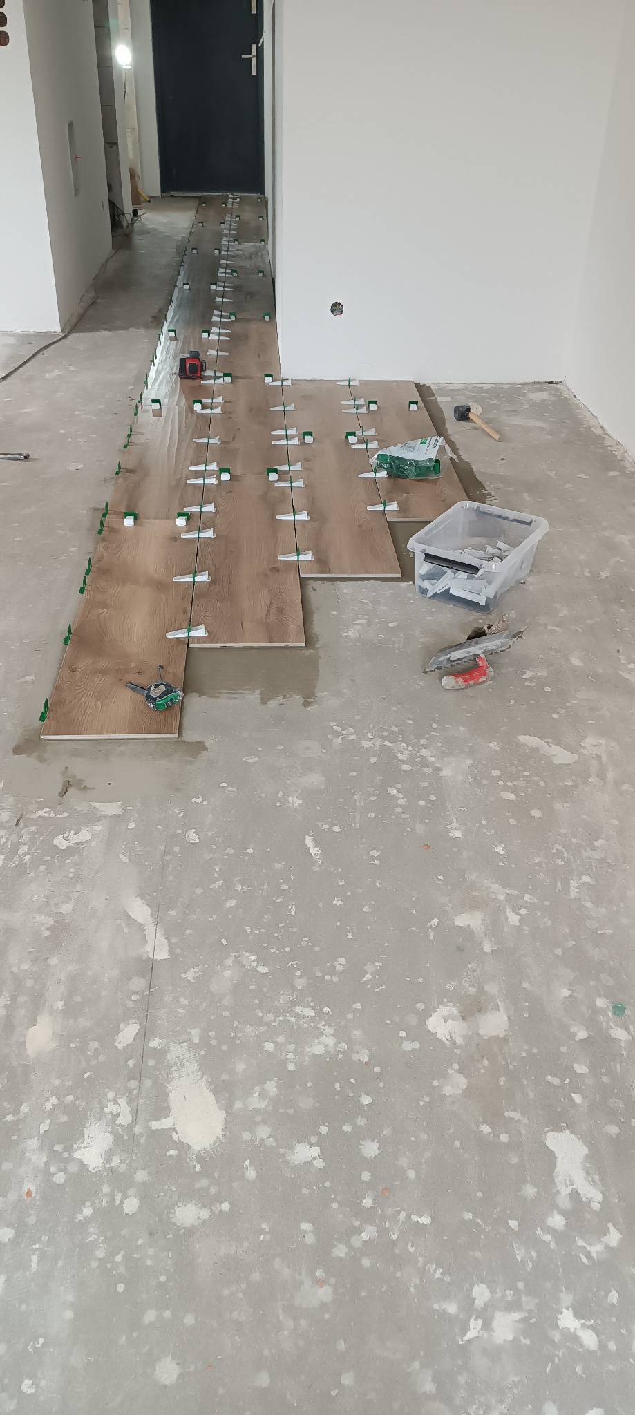 Układanie podłogi z paneli imitujących drewno w korytarzu, widoczne narzędzia glazurnicze i poziomica laserowa.