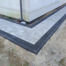 Tomasz Grzeszczak - Świeżo ułożony chodnik z szarej kostki brukowej z czarnym obramowaniem, przylegający do ściany budynku z widoczną rurą spustową, na surowym, niezagospodarowanym jeszcze gruncie.