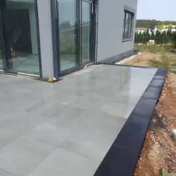 Nowoczesny taras z szarych płyt, obrzeża wykończone czarną kostką, widoczne narzędzia brukarskie przy ścianie budynku.