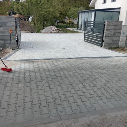 Toms-bruk - Nowoczesny podjazd z jasnoszarej kostki brukowej prowadzący do domu z szarą bramą. Widoczny fragment starszej nawierzchni i narzędzia brukarskie.