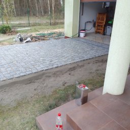 Toms-bruk - Ułożona kostka granitowa przed garażem, widoczny fragment podjazdu i przygotowane podłoże. W tle las i elementy wyposażenia garażu.