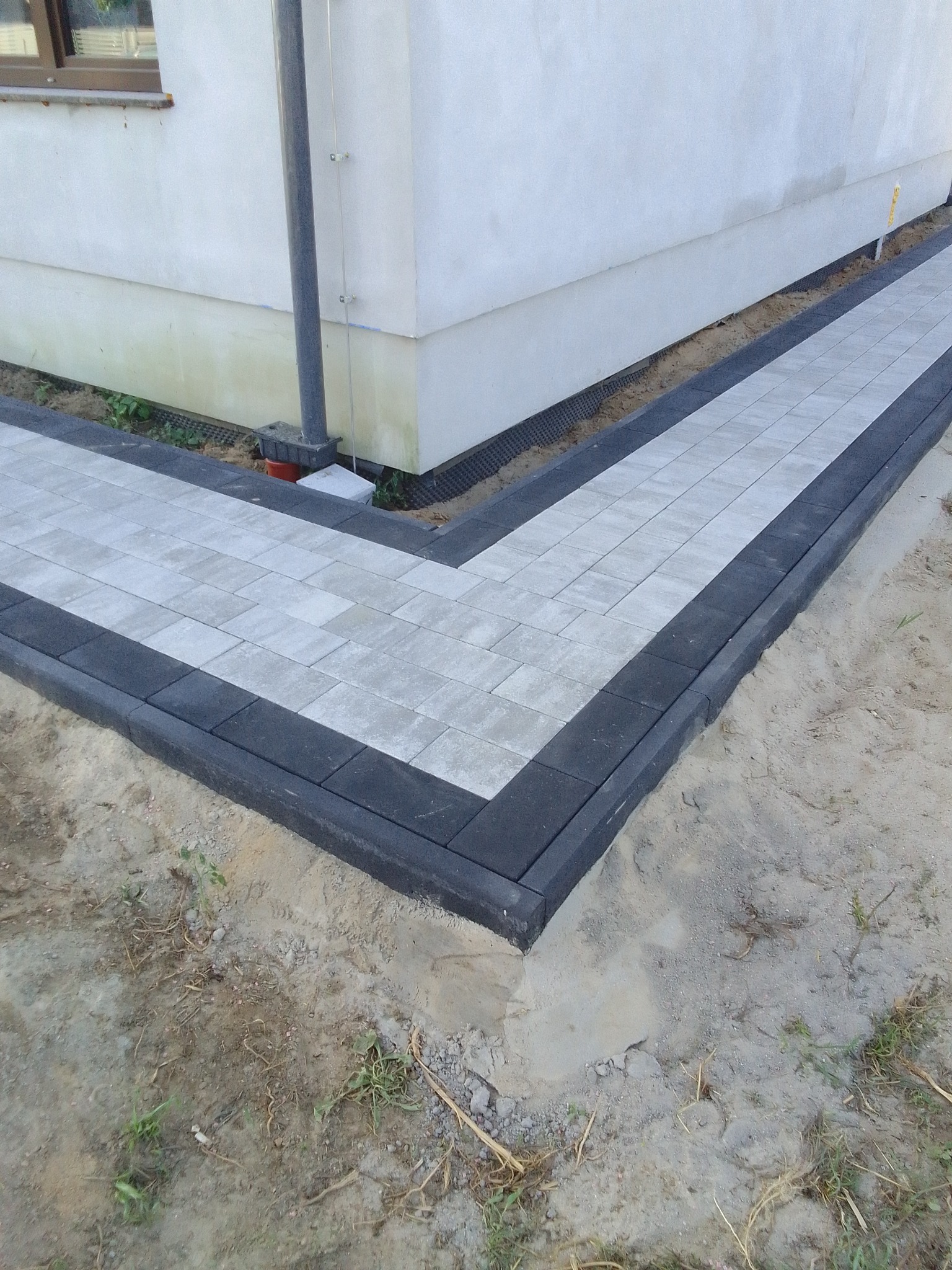 Świeżo ułożony chodnik z szarej kostki brukowej z czarnym obramowaniem, przylegający do ściany budynku z widoczną rurą spustową, na surowym, niezagospodarowanym jeszcze gruncie.