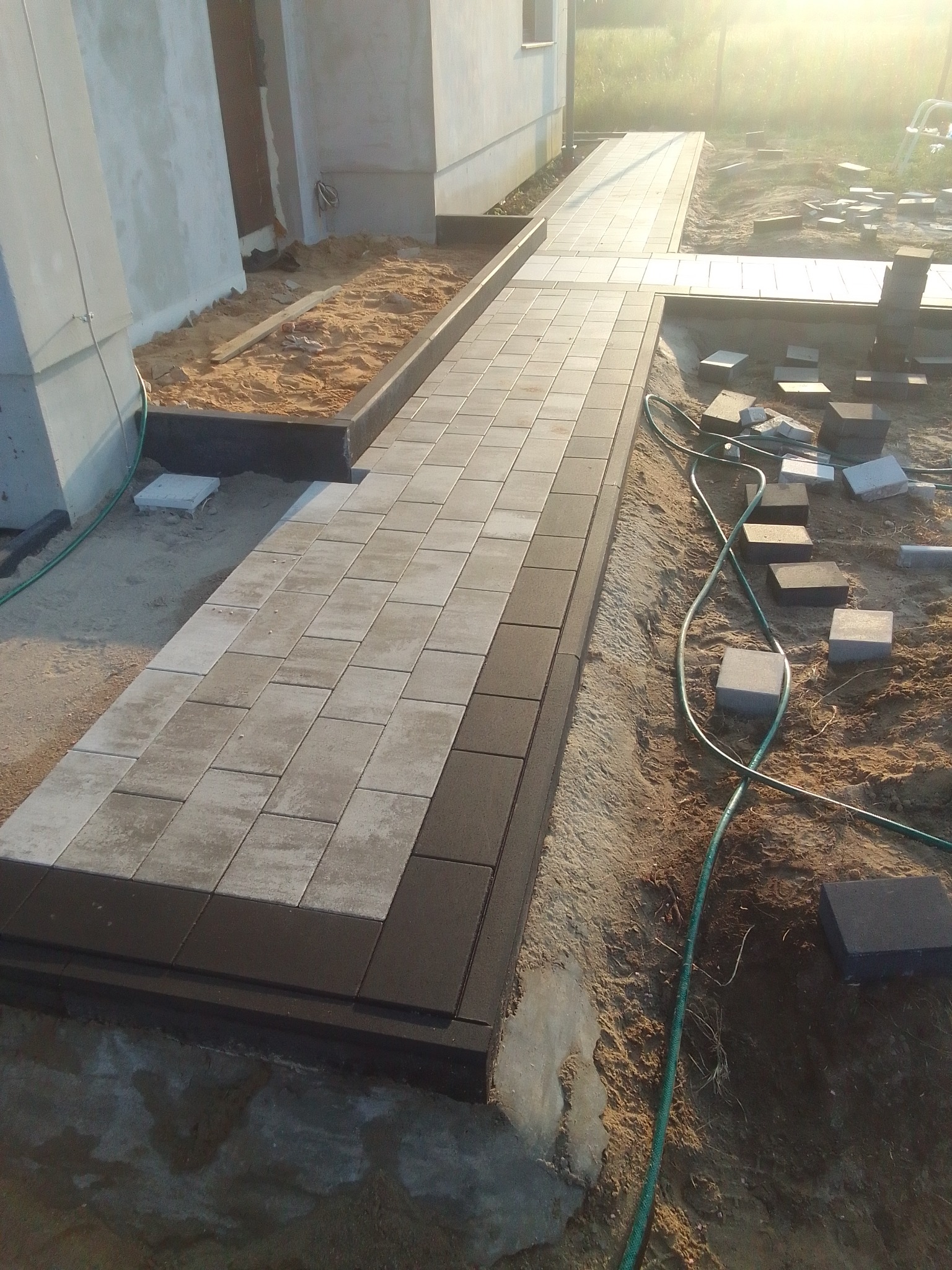 Ścieżka z szarej kostki brukowej z ciemnymi obrzeżami prowadząca wzdłuż budynku, widoczne luźne kostki i wąż ogrodowy na piasku.