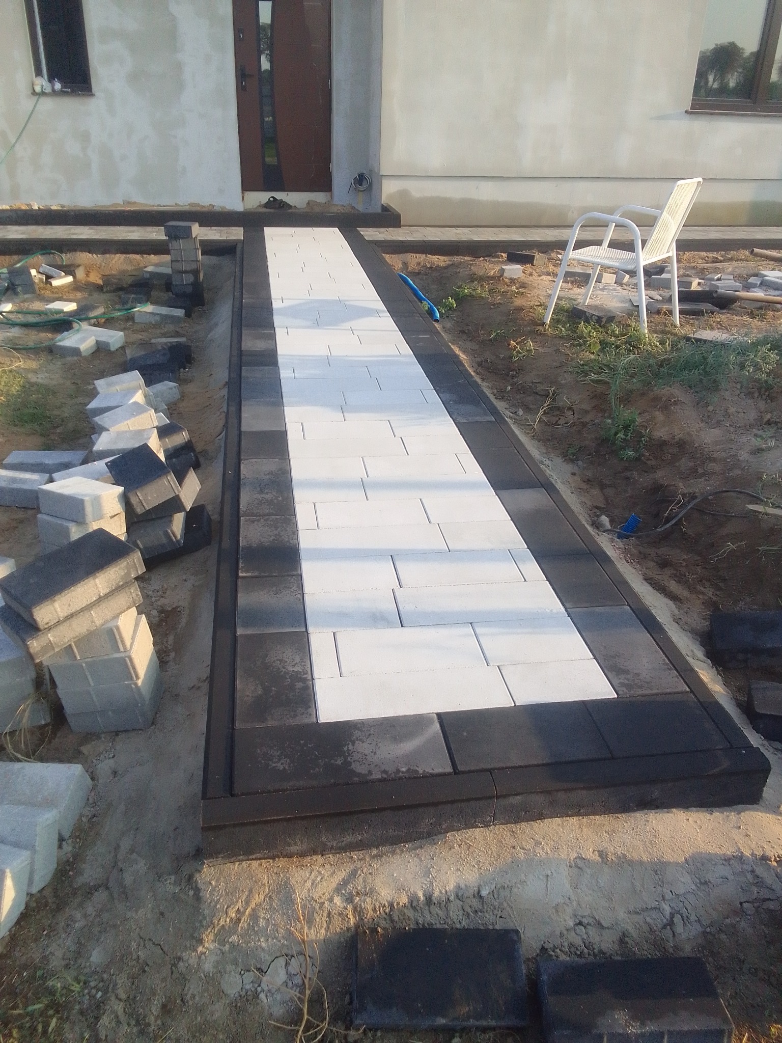 Ścieżka z jasnej kostki brukowej z ciemną obwódką w trakcie układania, widoczne stosy kostki i białe krzesło ogrodowe w tle.