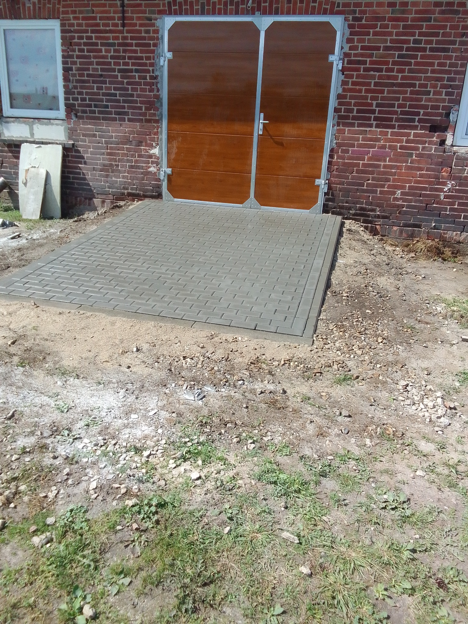Ułożony chodnik z szarej kostki brukowej prowadzący do brązowych drzwi garażowych w budynku z czerwonej cegły, widoczny piasek i gruz wokół.