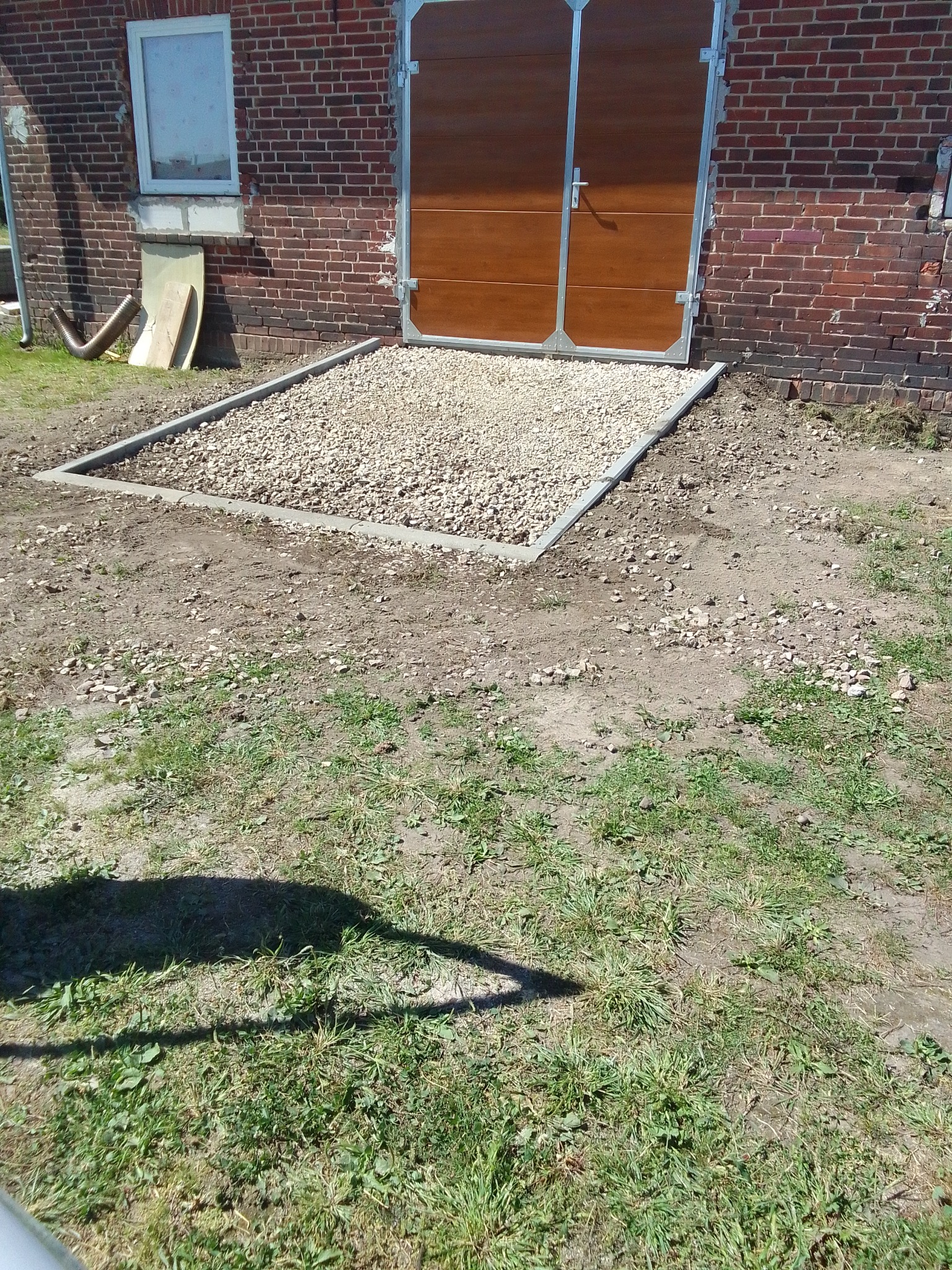 Przygotowany teren przed budynkiem z cegły, wyznaczony betonową obwódką i wypełniony żwirem, prowadzący do dwuskrzydłowych, brązowych drzwi garażowych.