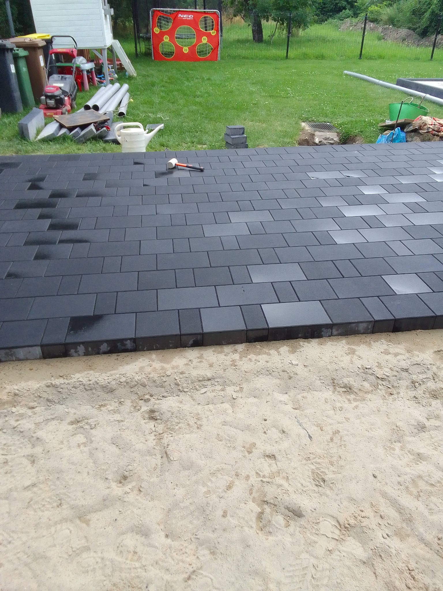 Częściowo ułożona nawierzchnia z ciemnoszarej kostki brukowej na podsypce piaskowej, z gumowym młotkiem leżącym na kostce, w tle trawnik z narzędziami i sprzętem ogrodowym.