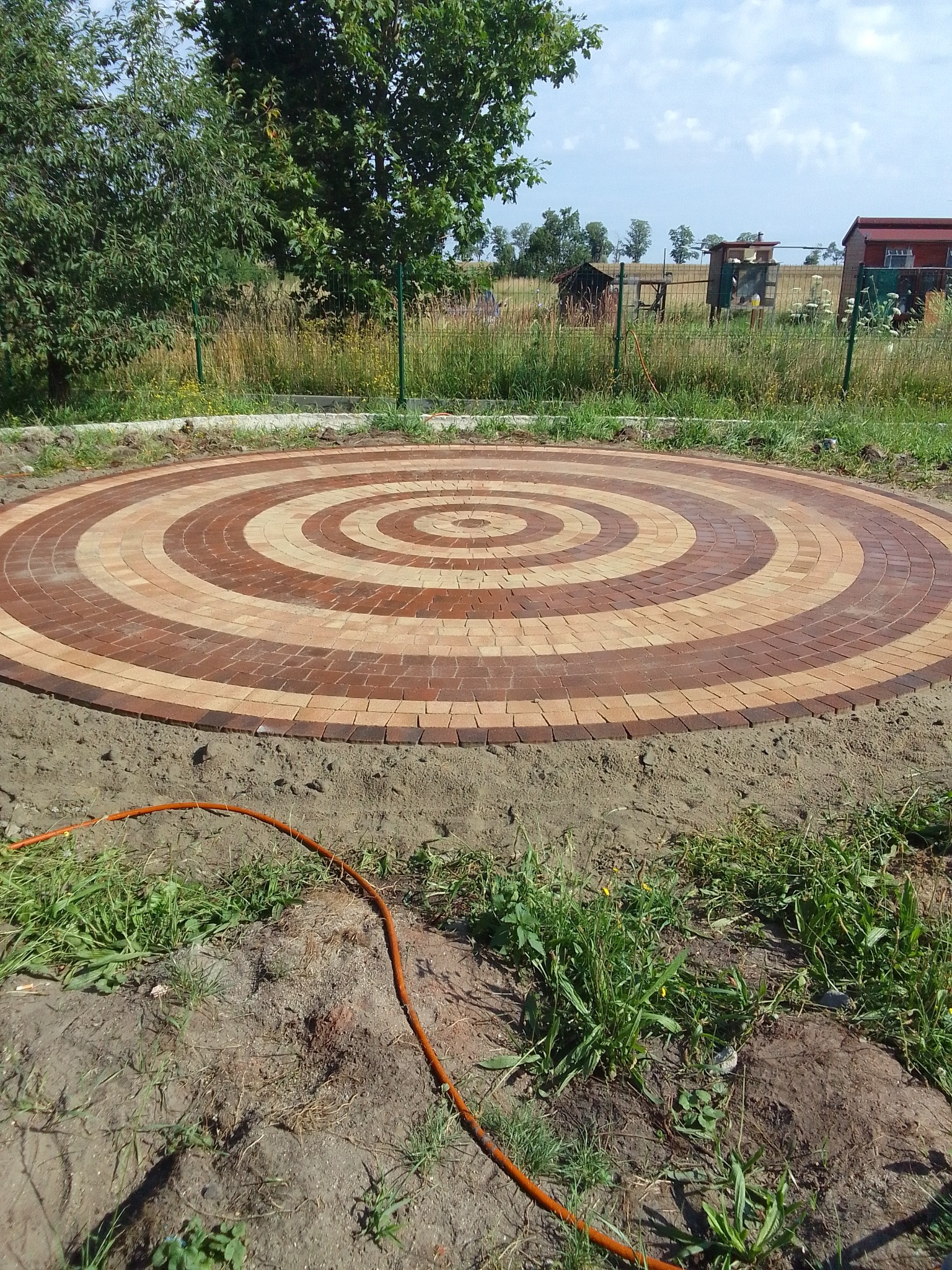 Okrągły plac z kostki brukowej ułożonej w koncentryczne kręgi z brązowej i beżowej kostki, widok z góry, w tle zieleń i zabudowania gospodarcze.