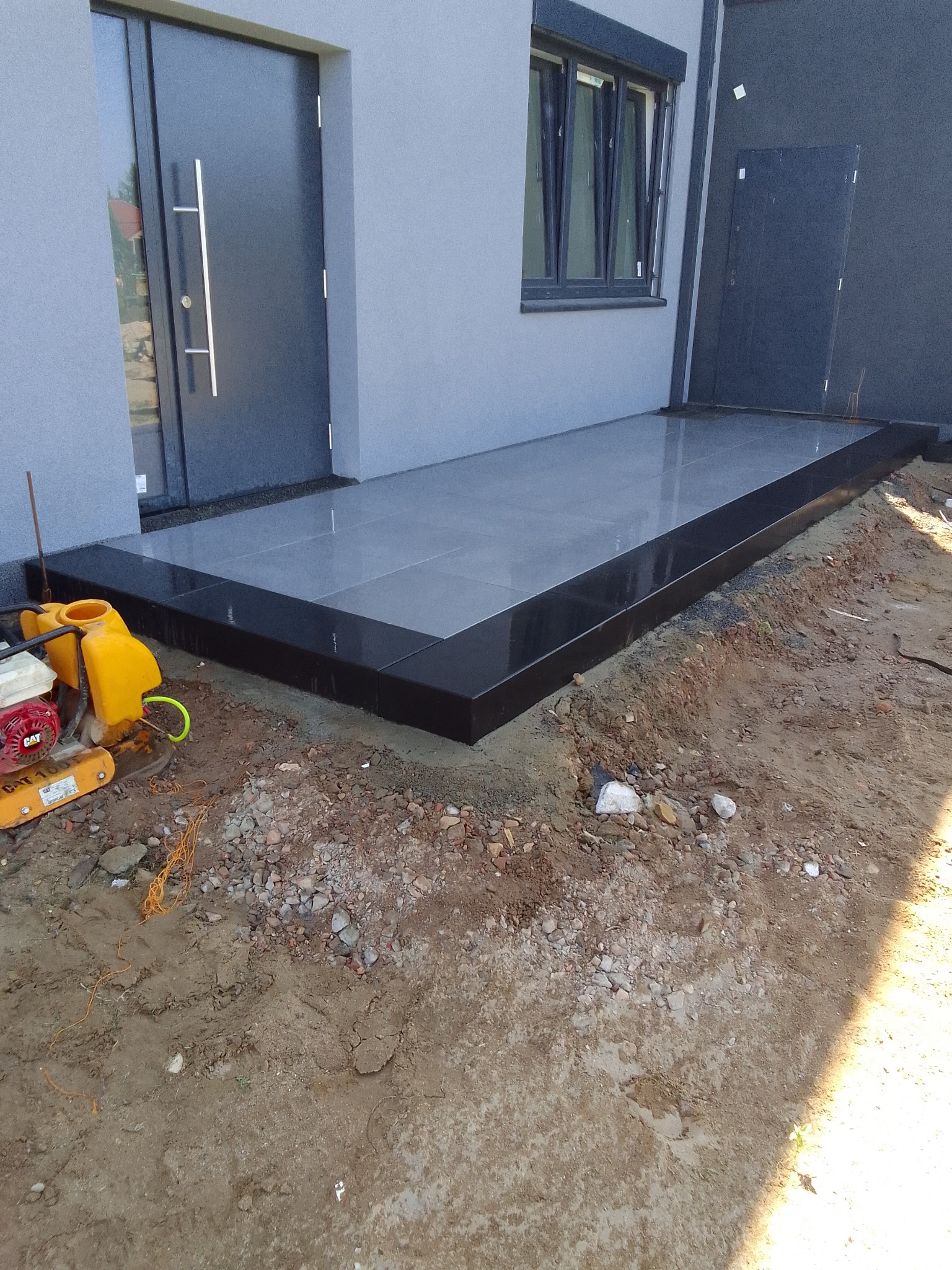 Wykończony, nowoczesny taras przed domem z szarych płytek z czarną obwódką, obok stoi zagęszczarka w kolorze żółtym, teren wokół jeszcze niezagospodarowany.