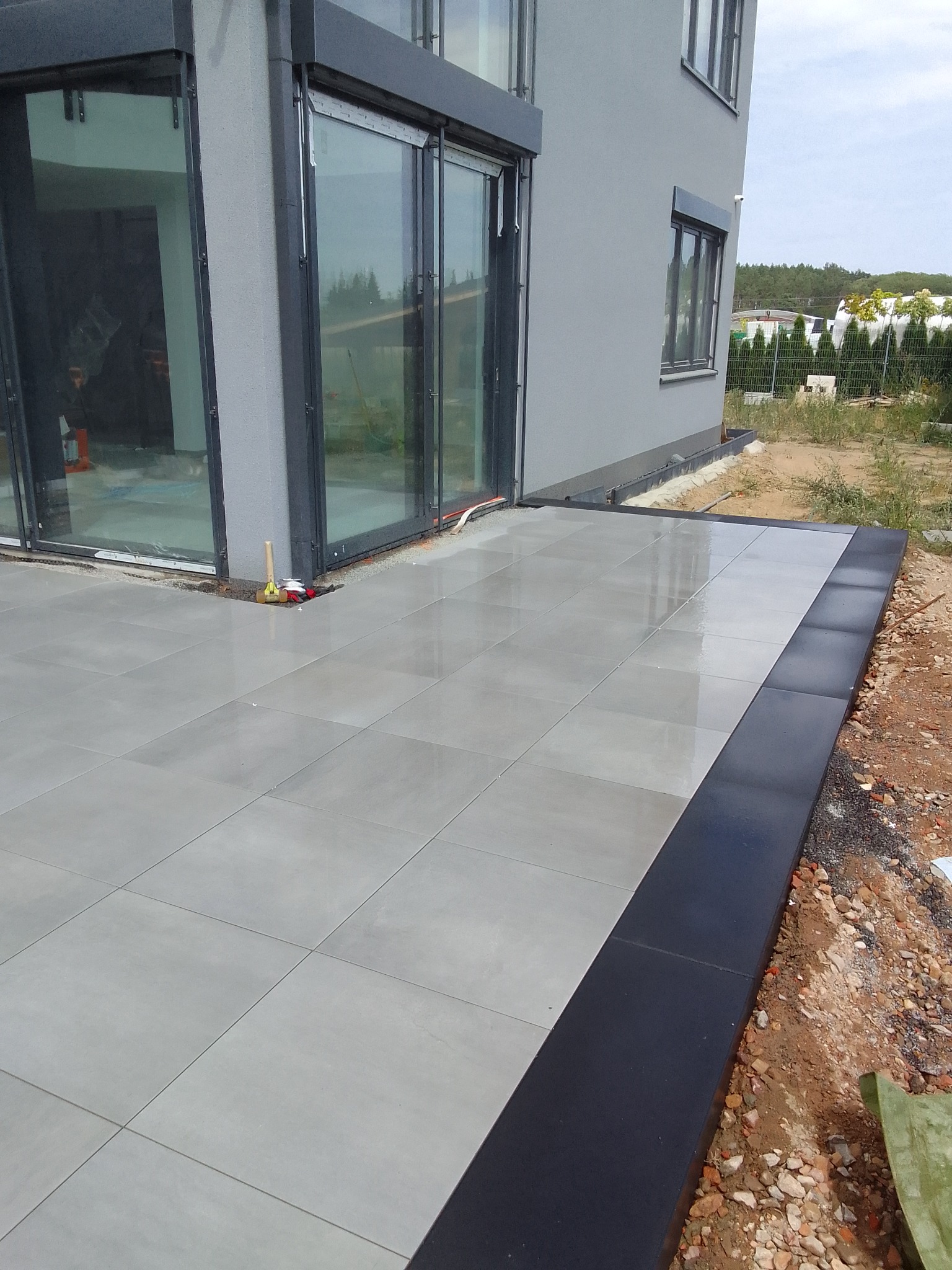 Nowoczesny taras z szarych płyt, obrzeża wykończone czarną kostką, widoczne narzędzia brukarskie przy ścianie budynku.