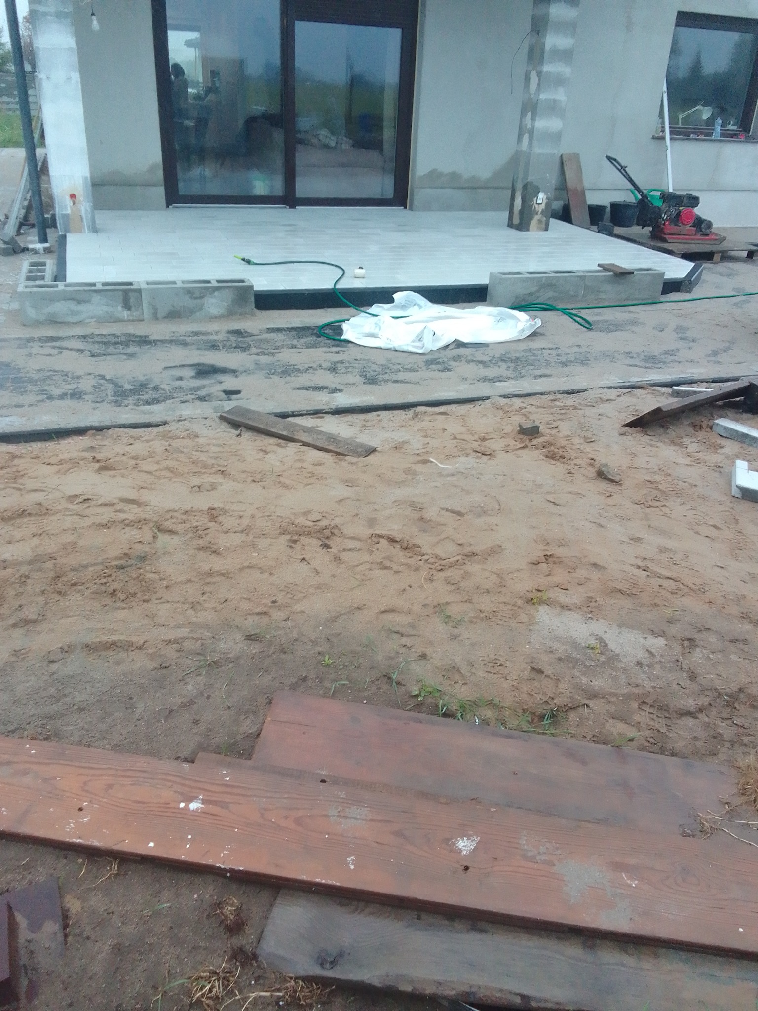 Przygotowanie terenu pod taras z kostki: piasek, deski, białe bloczki betonowe, ubijarka spalinowa i częściowo ułożona szara kostka przed budynkiem.