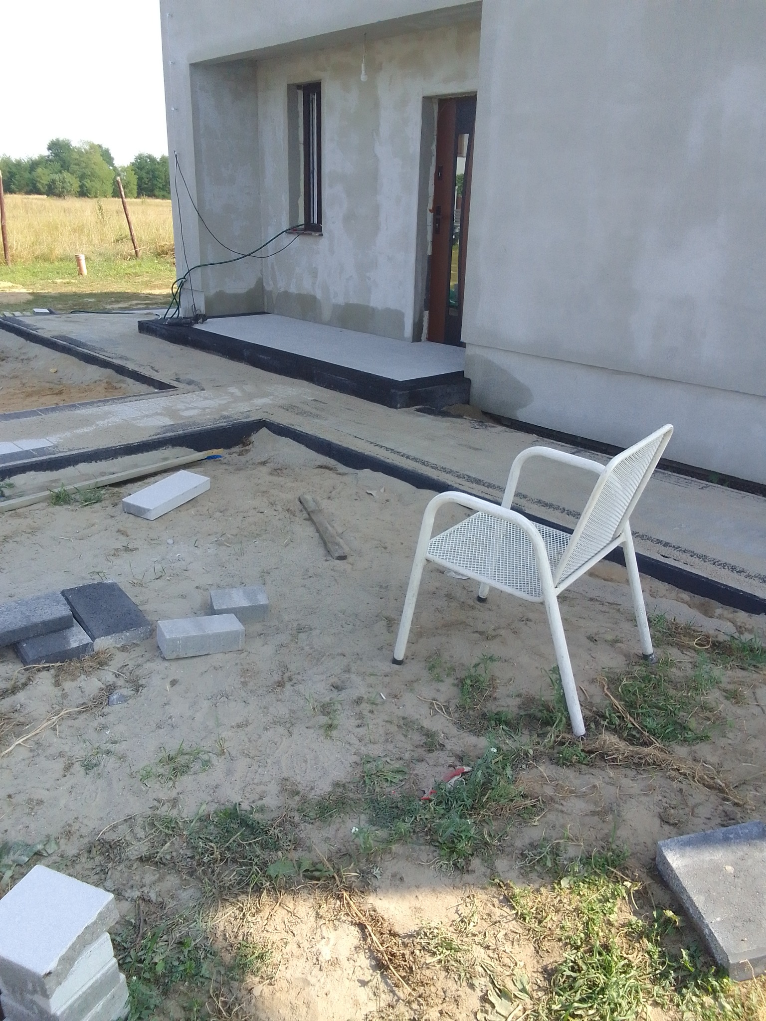 Teren przed nowym domem w trakcie przygotowywania podłoża pod nawierzchnię, widoczne obrzeża, piasek, ułożone płyty chodnikowe i krzesło ogrodowe.