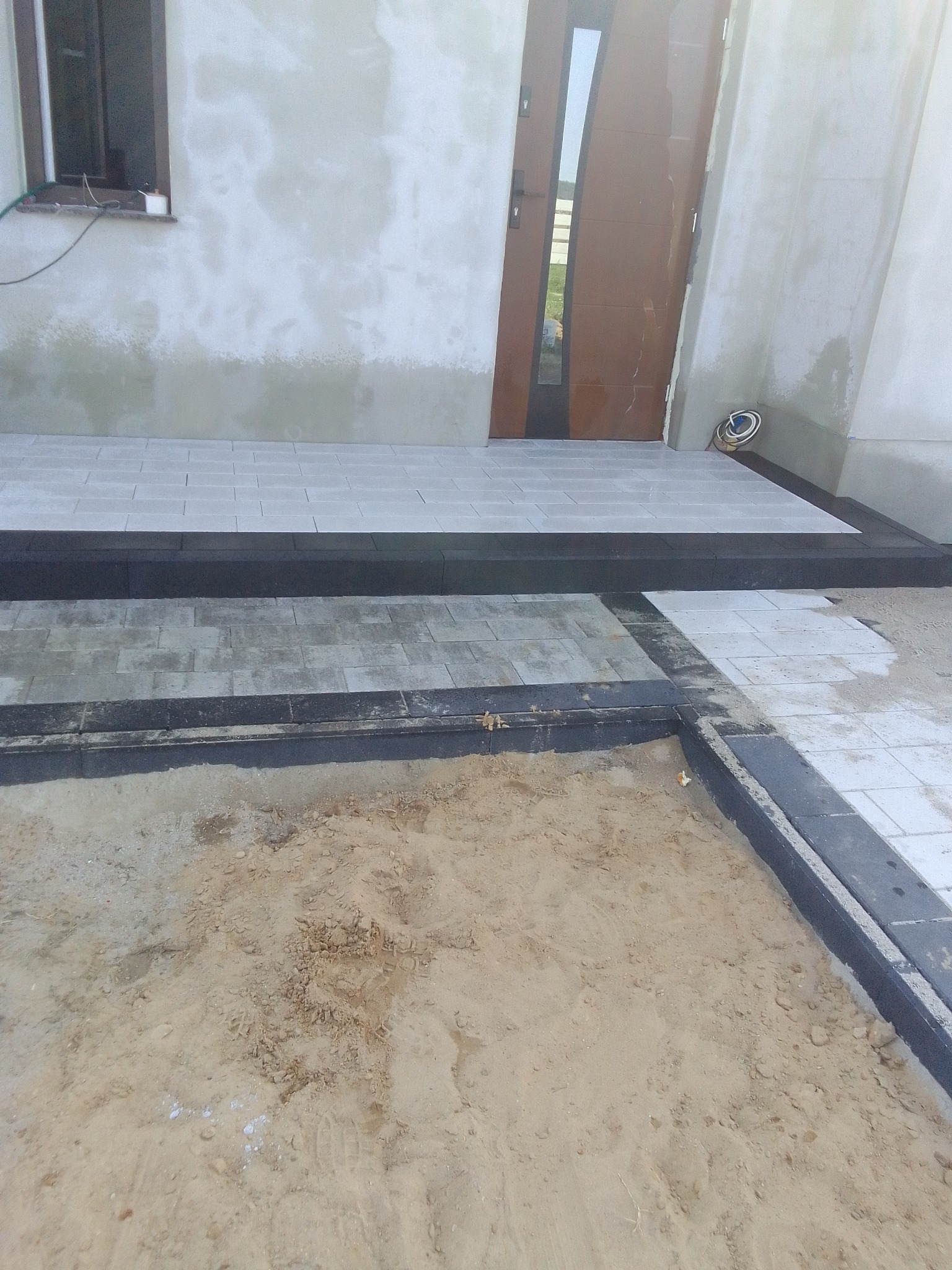Fragment układania jasnoszarej kostki brukowej przed wejściem do domu, widoczny piasek i obrzeża z ciemnej kostki.