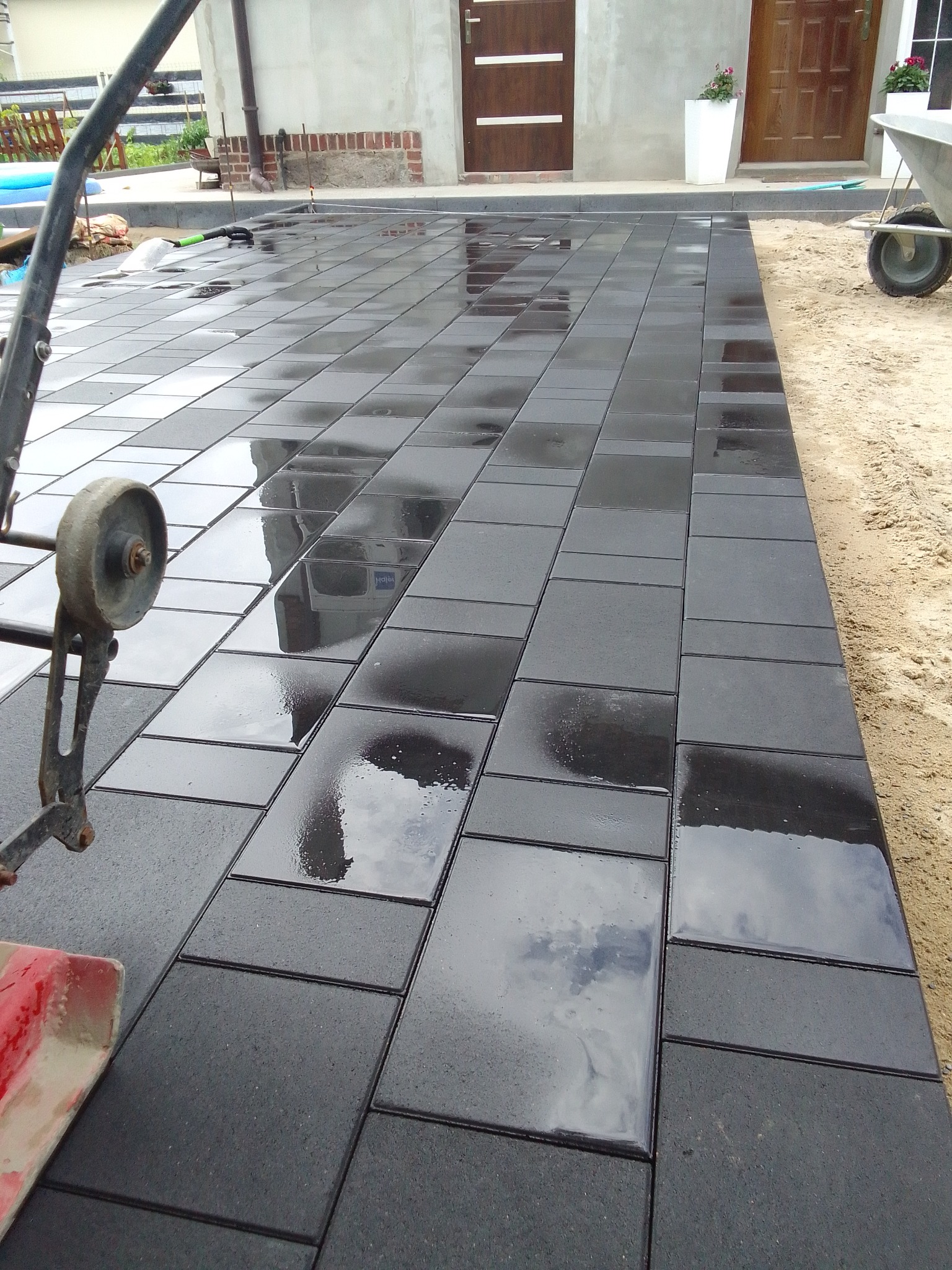 Świeżo ułożona, mokra, ciemnoszara kostka brukowa w regularnym wzorze, z widocznymi refleksami nieba i budynków, obok piasek i narzędzia brukarskie.