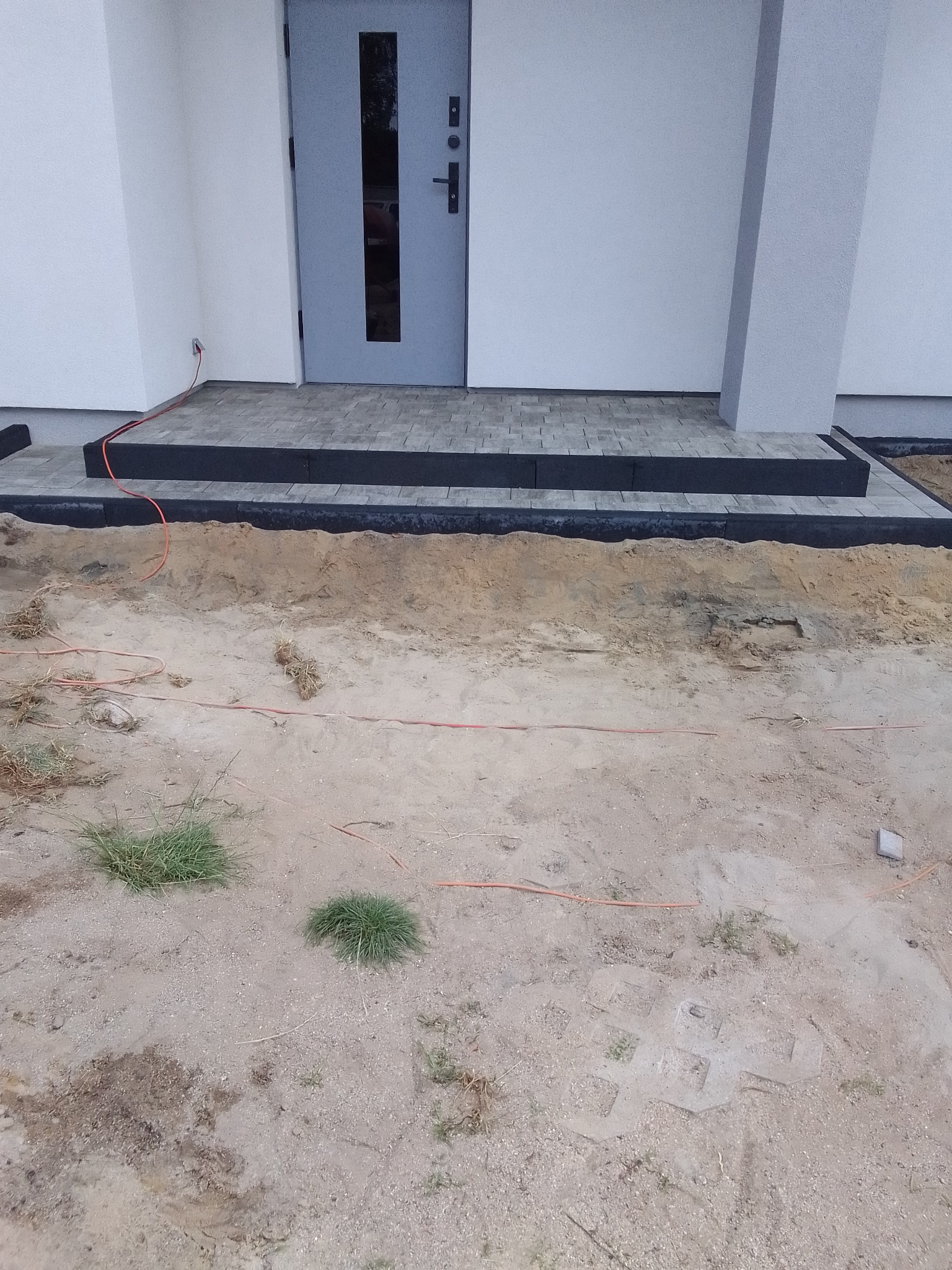 Schody wejściowe do domu z kostki brukowej, widoczny kabel elektryczny i teren przed domem w trakcie przygotowania.