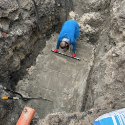 Profi Trax Rafał Jeglorz - Pracownik w niebieskiej bluzie i czerwonych rękawicach poziomuje betonową płytę w wykopie za pomocą poziomicy; widoczne narzędzia, rura PCV i worek cementu.