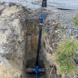 Głęboki wykop w ziemi z widocznymi rurami wodociągowymi z niebieskimi zaworami, zabezpieczony taśmą ostrzegawczą, z elementami instalacji wystającymi ponad powierzchnię gruntu.