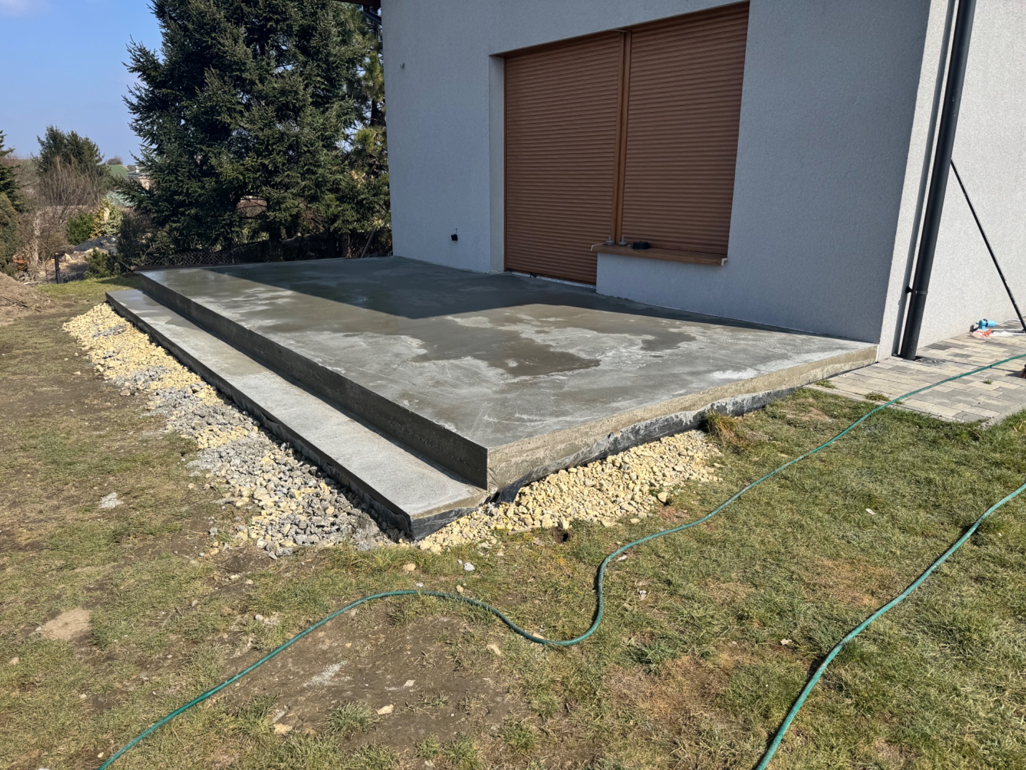 Świeżo wylany betonowy taras z trzema stopniami przy budynku z brązowymi roletami, otoczony kamieniami i trawnikiem, widoczny wąż ogrodowy.