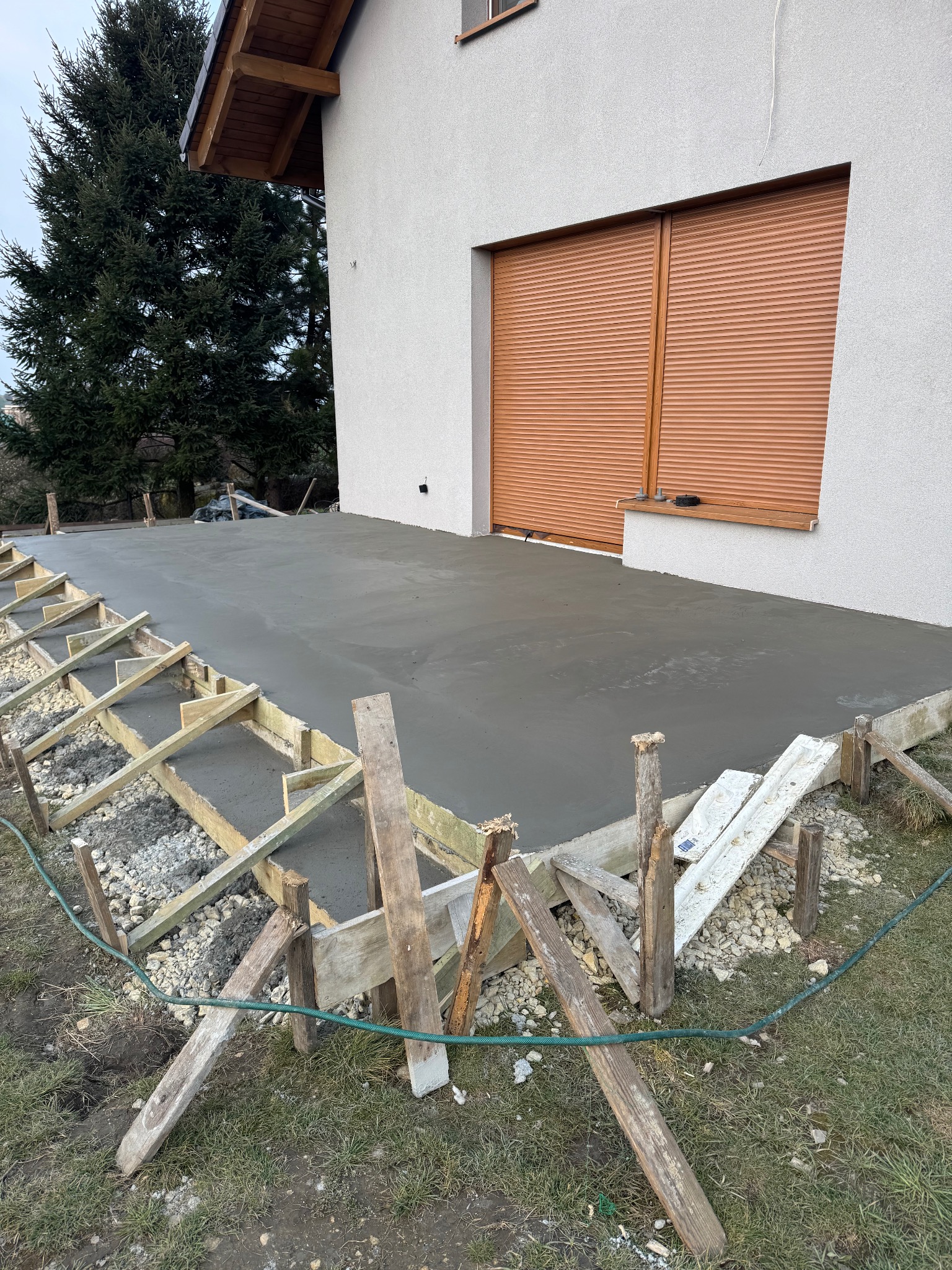 Wylewka betonowa na przygotowanym podłożu z drewnianym szalunkiem przy ścianie budynku z brązowymi roletami.