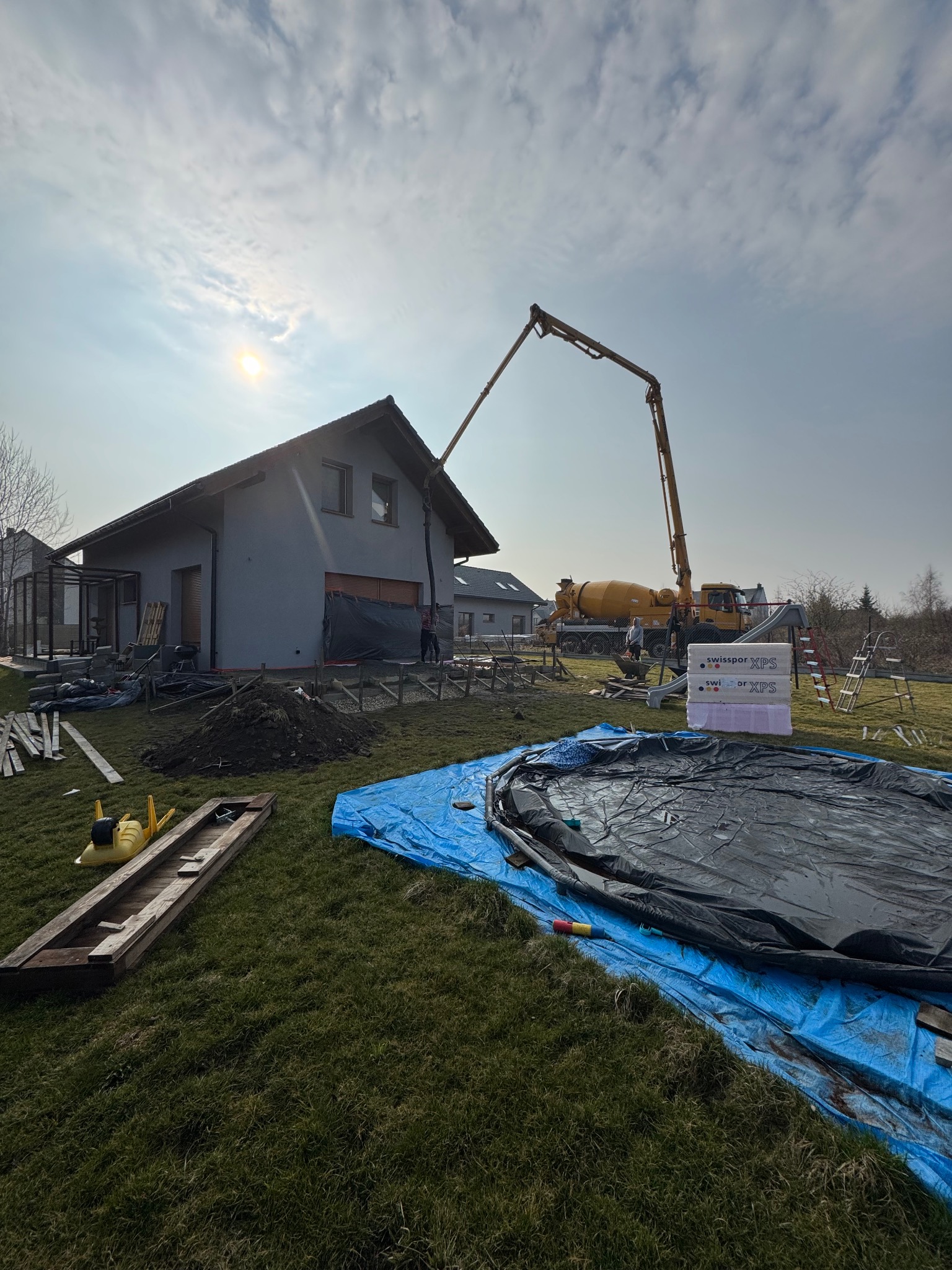 Wylewka betonu do fundamentów domu jednorodzinnego przy użyciu pompy do betonu, widoczne szalunki, izolacja termiczna, przygotowany teren pod budowę.