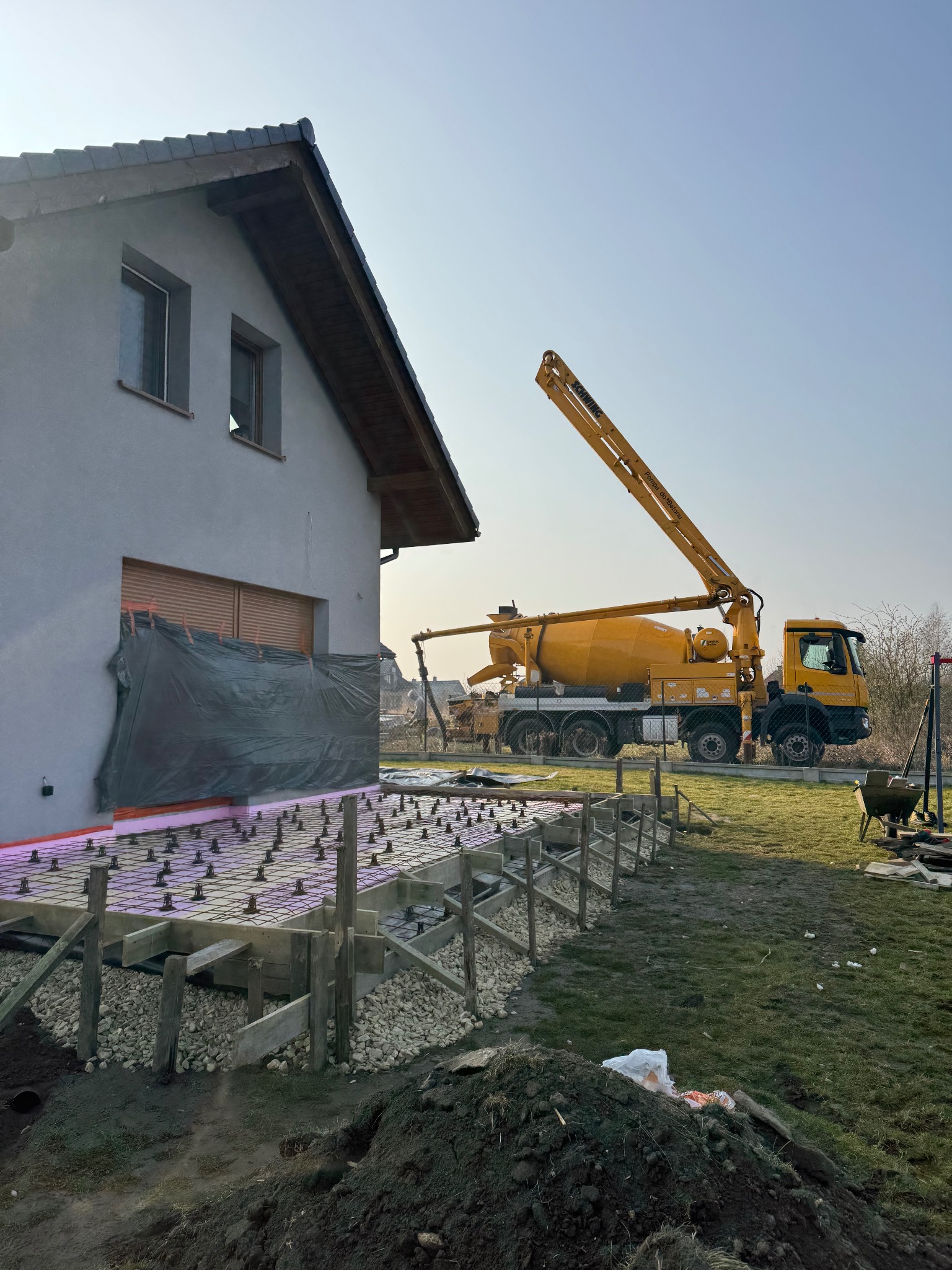 Przygotowanie fundamentów pod budynek jednorodzinny: szalunek z desek, izolacja termiczna z różowego styropianu na podłożu z folii, stalowe pręty zbrojeniowe i żółta betoniarka z wysięgnikiem w tle.