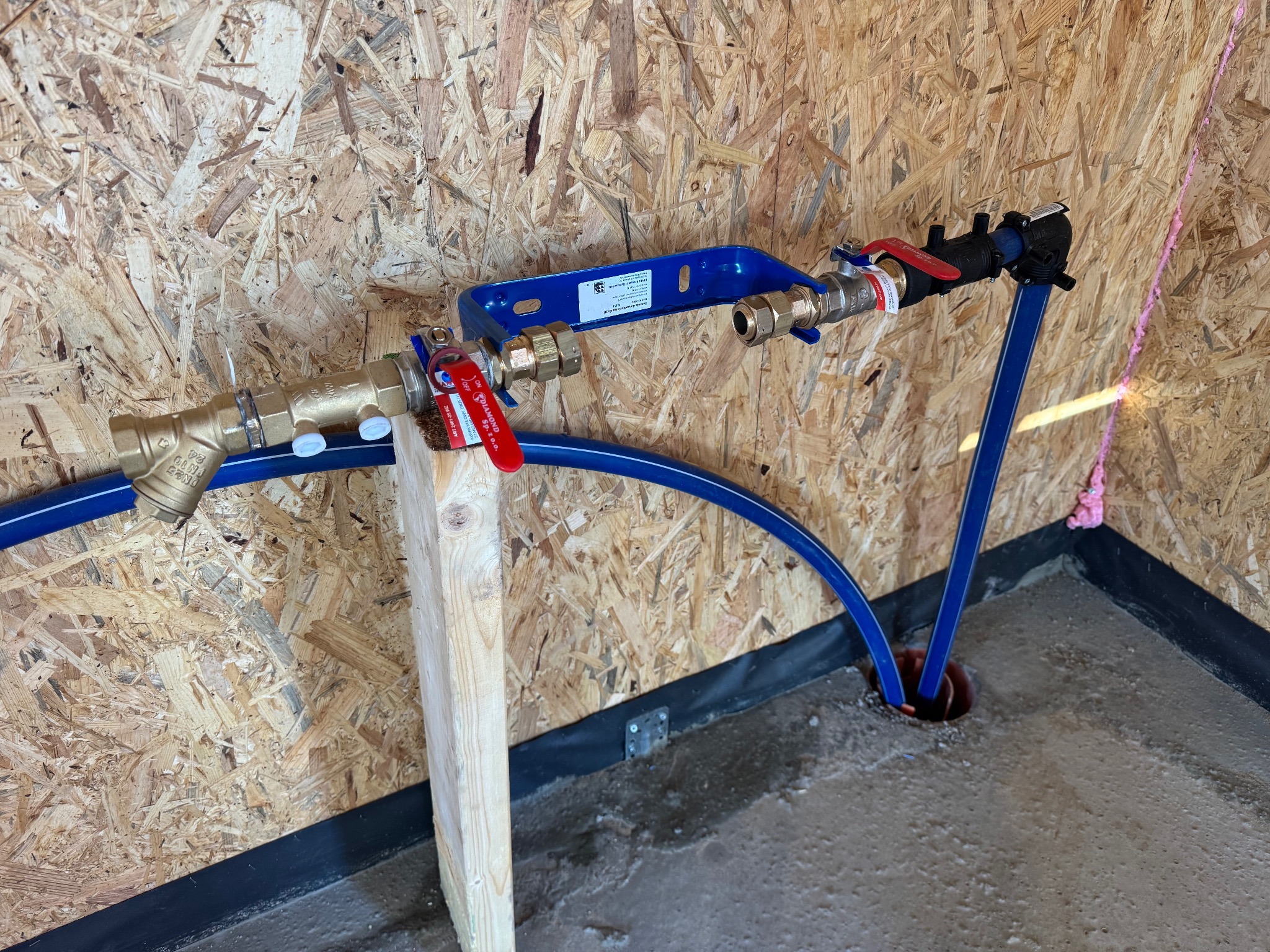 Fragment instalacji wodnej z niebieskimi rurami PEX, mosiężnymi zaworami i niebieskim uchwytem mocującym, zamontowanej na ścianie z płyty OSB i podłodze z wylewki betonowej. Widoczny drewniany...