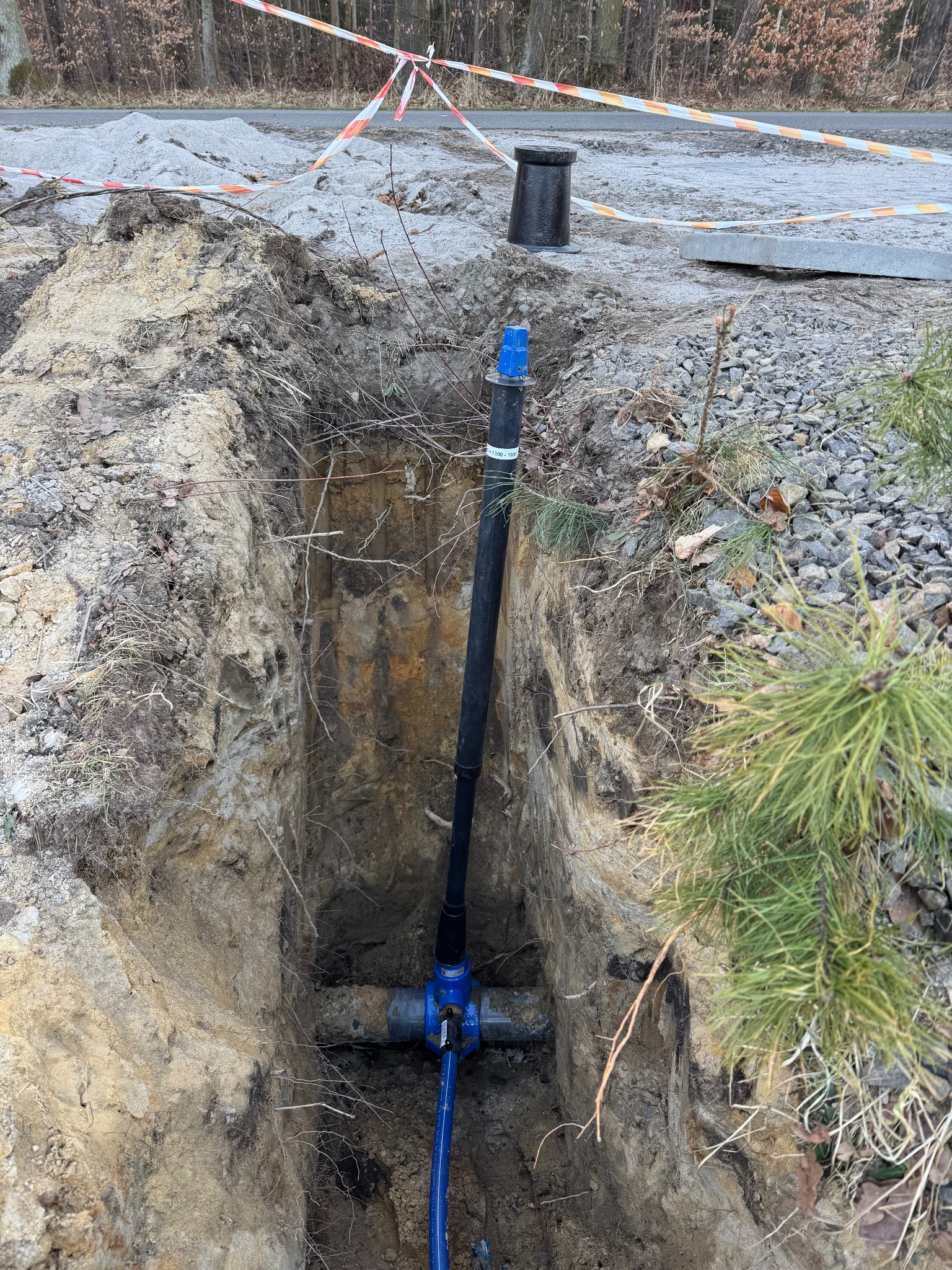 Głęboki wykop w ziemi z widocznymi rurami wodociągowymi z niebieskimi zaworami, zabezpieczony taśmą ostrzegawczą, z elementami instalacji wystającymi ponad powierzchnię gruntu.