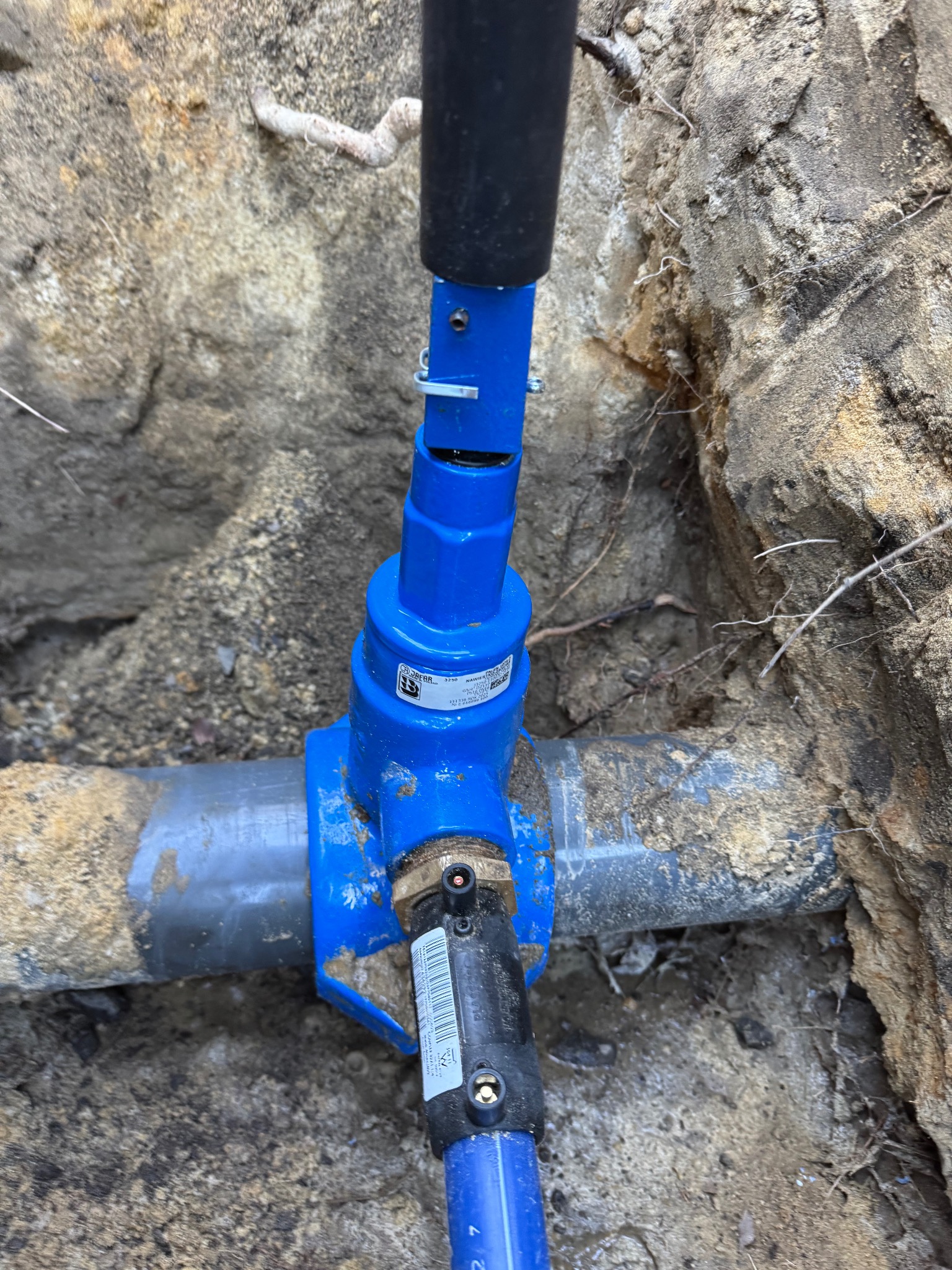 Niebieski zawór na szarej rurze w ziemi, widoczne ślady świeżego wykopu i korzenie