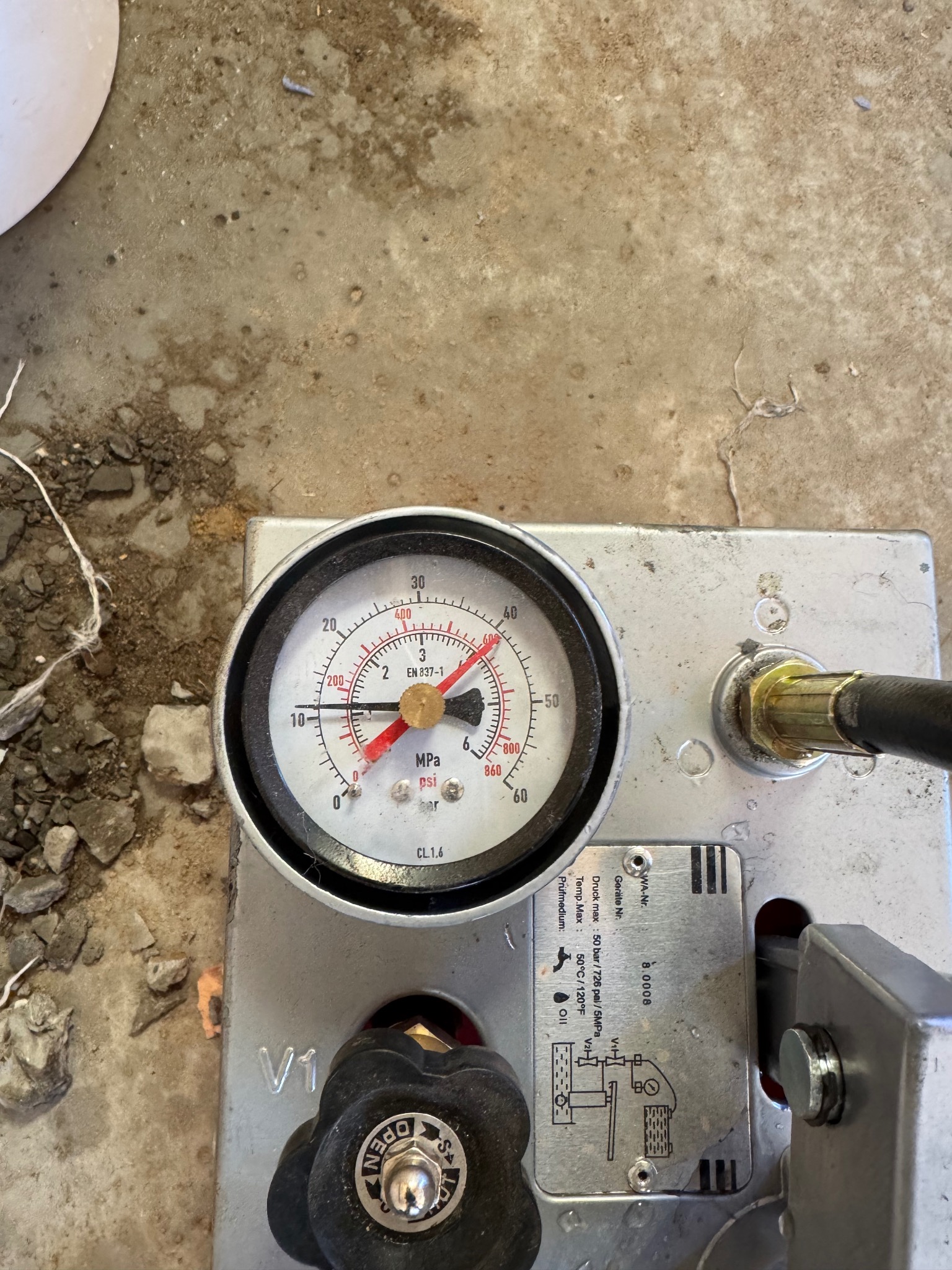 Manometr ciśnieniowy, wskazujący niskie ciśnienie, na metalowej podstawie z widocznym schematem hydraulicznym i pokrętłem, leżący na betonowej powierzchni z drobnymi kamieniami.