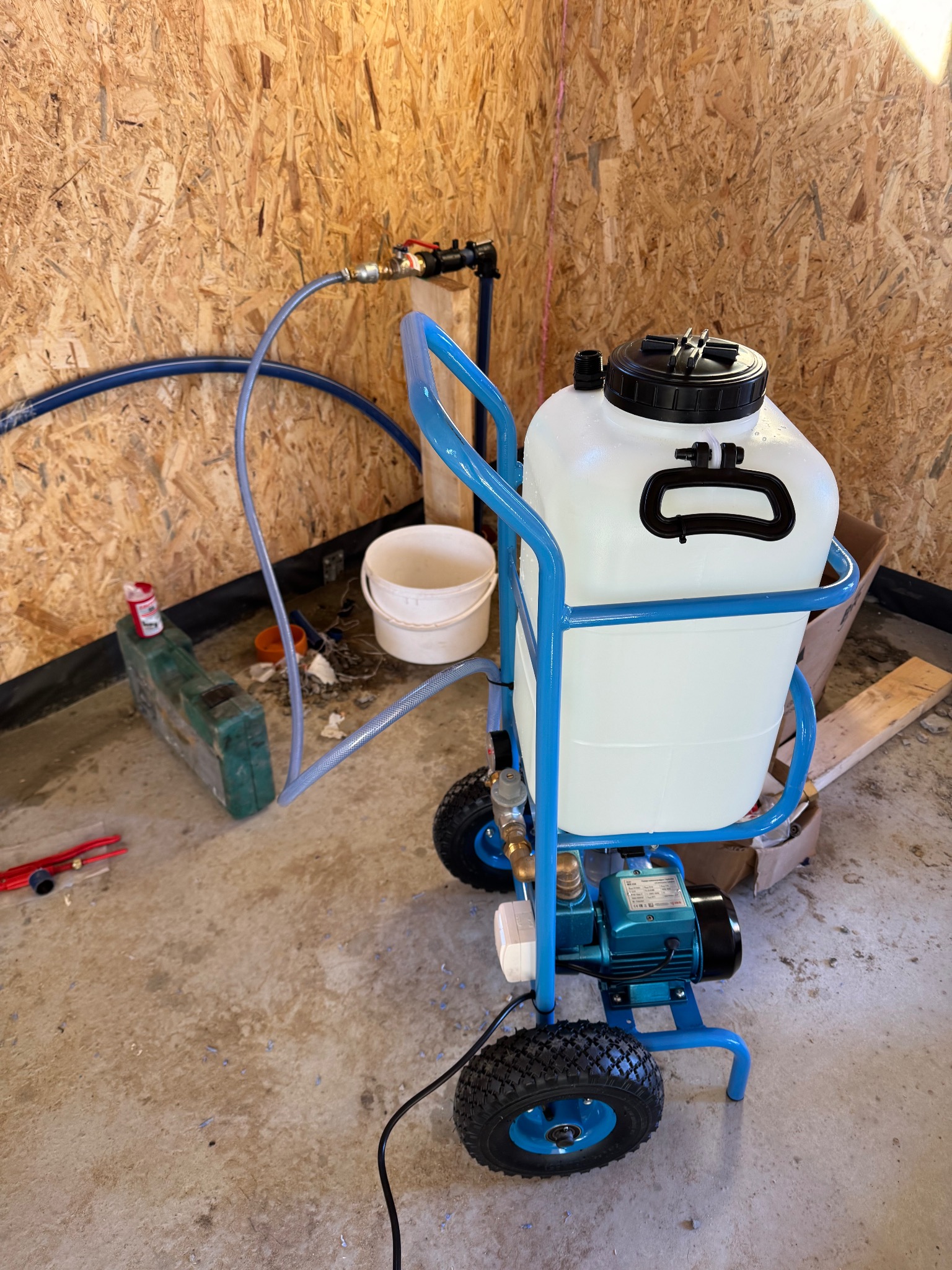 Niebieski wózek z białym zbiornikiem i pompą elektryczną, podłączony do instalacji wodnej w pomieszczeniu wyłożonym płytami OSB.