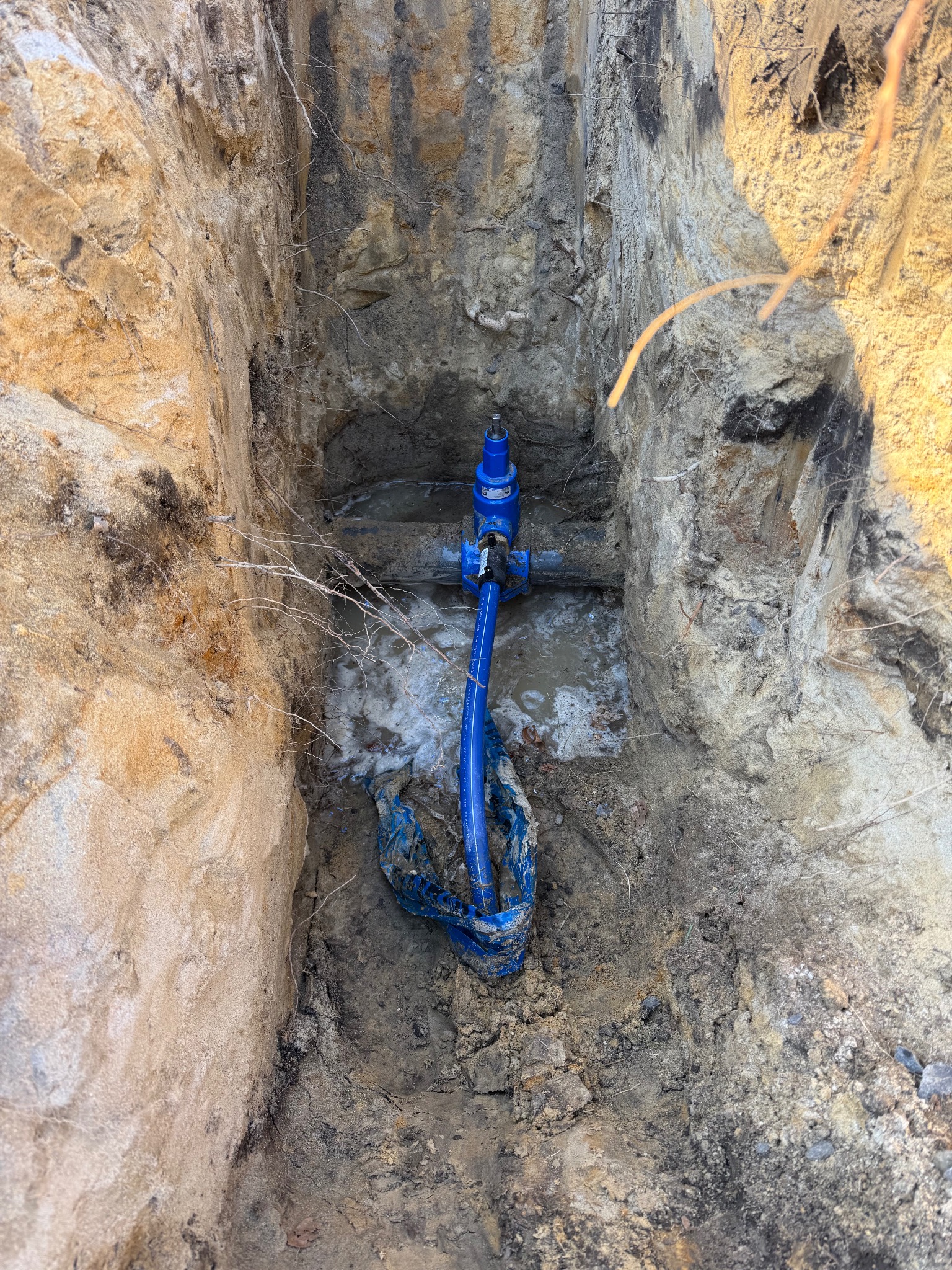 Wykop w ziemi z widocznym, częściowo zamontowanym, niebieskim zaworem i rurami wodociągowymi, na dnie wykopu woda i kawałek niebieskiej folii.