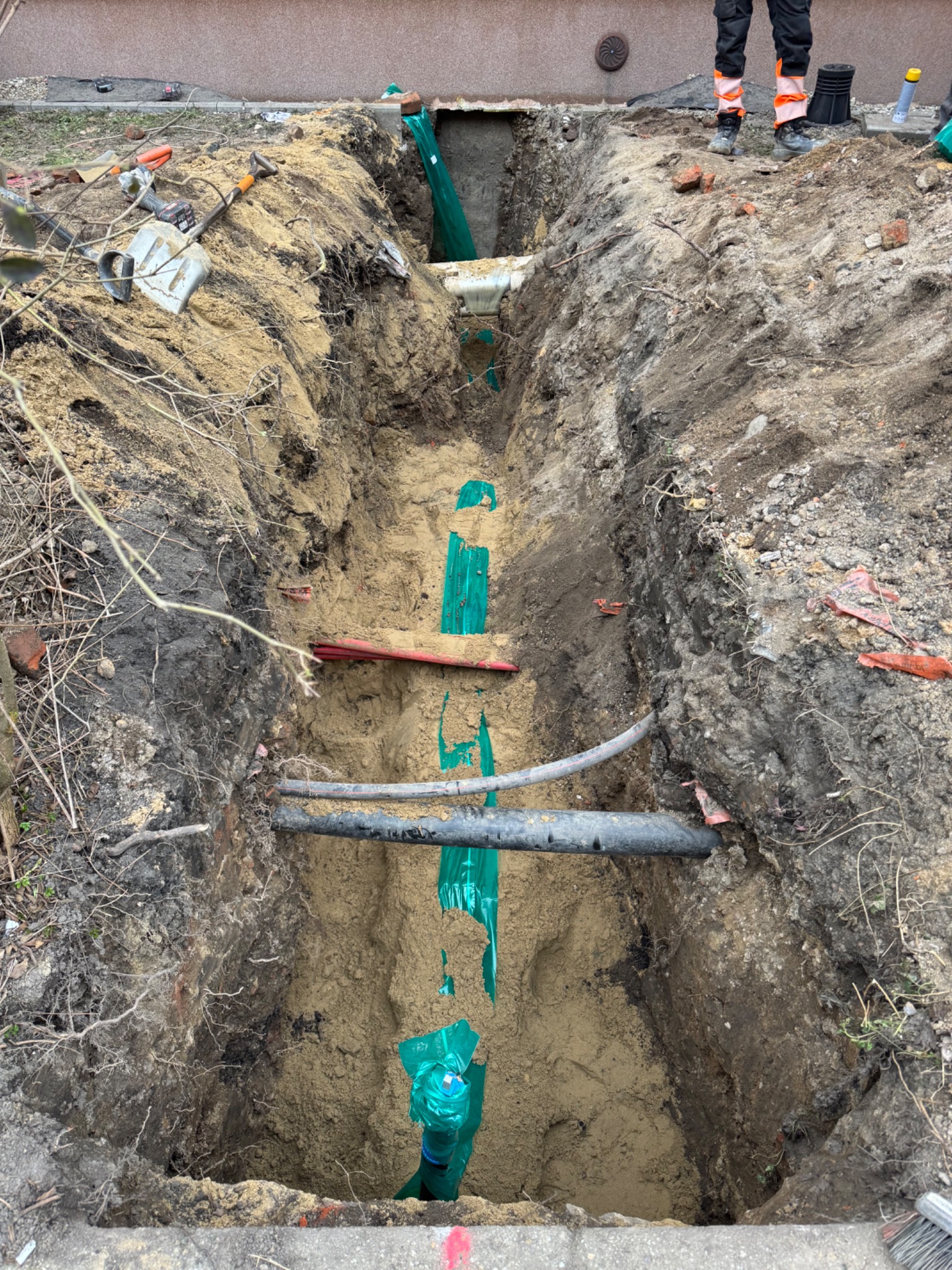Głęboki wykop w ziemi z widocznymi rurami, zabezpieczonymi zieloną folią, narzędzia budowlane i resztki cegieł na krawędzi wykopu, fragment osoby w odzieży roboczej w tle.