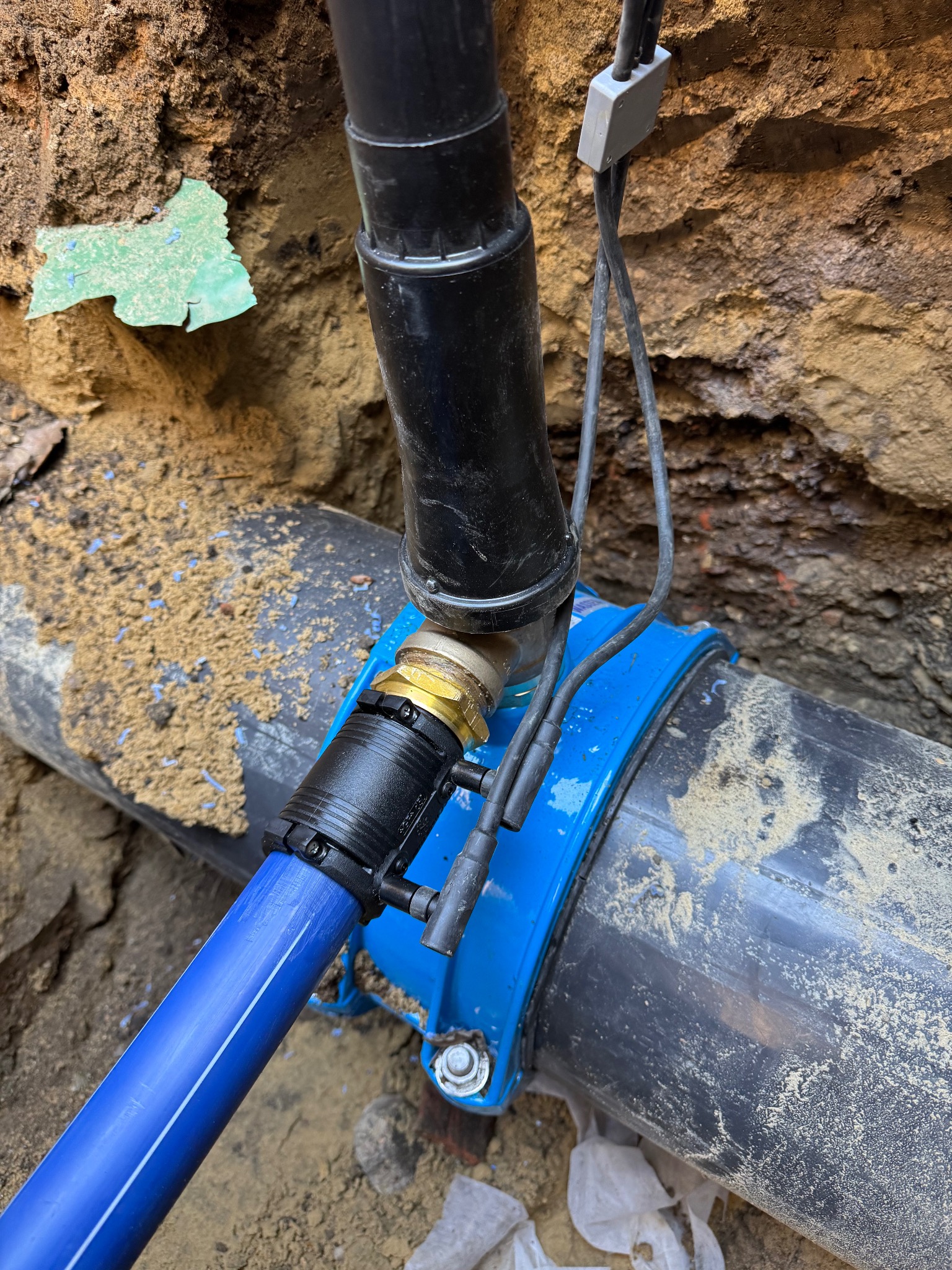 Połączenie rur: czarna rura pionowa łączy się z szarą rurą poziomą za pomocą niebieskiego zacisku i mosiężnej złączki, a niebieska rura odchodzi w lewo, całość w wykopie ziemnym.