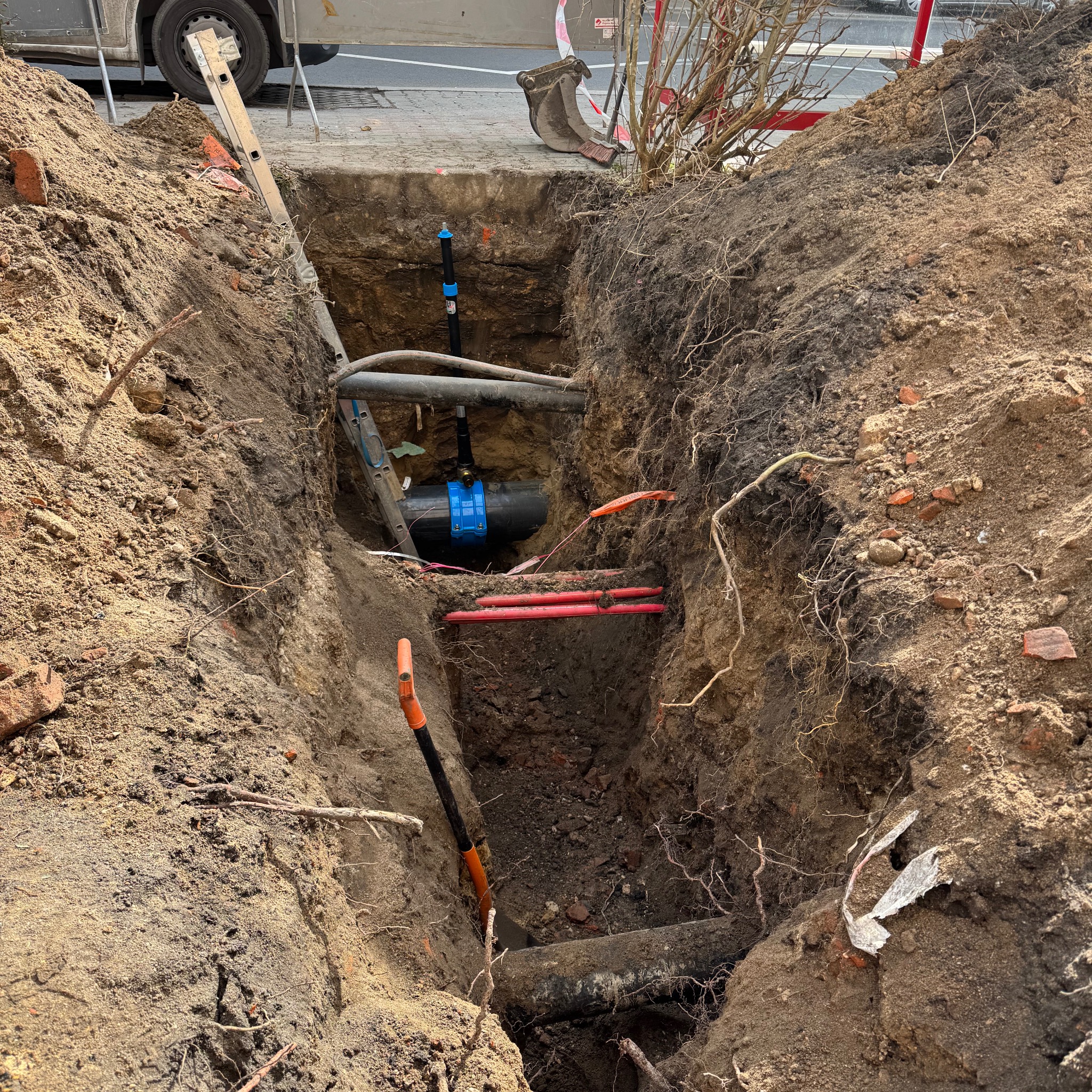 Głęboki wykop w ziemi z widocznymi rurami wodociągowymi, rurami osłonowymi kabli elektrycznych, oraz elementami instalacji kanalizacyjnej, z drabinką opartą o brzeg wykopu i łopatą wbitą w ziemię...