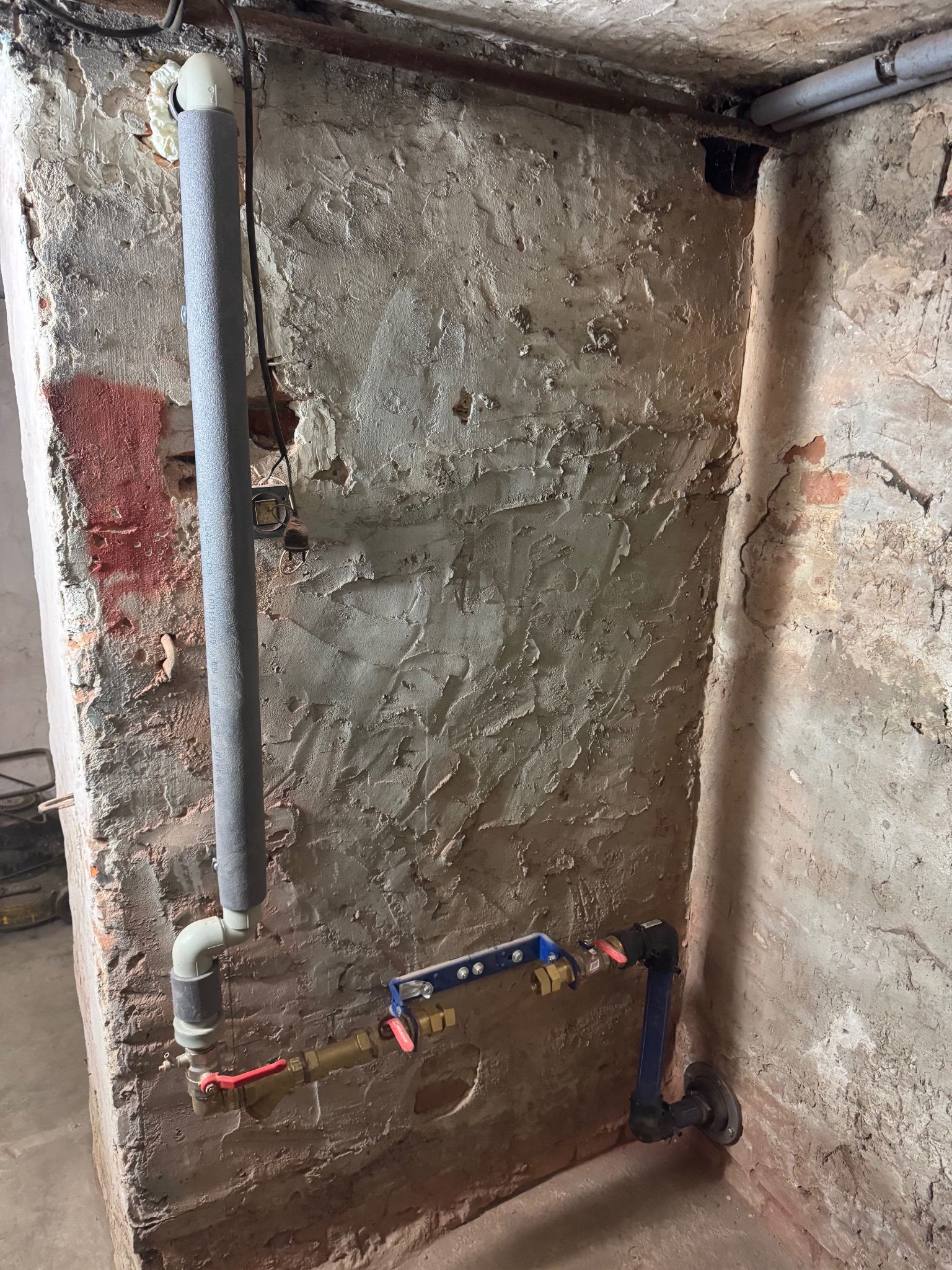 Instalacja wodna z zaworami i rurami z izolacją termiczną, zamontowana w rogu pomieszczenia o surowych, otynkowanych ścianach.