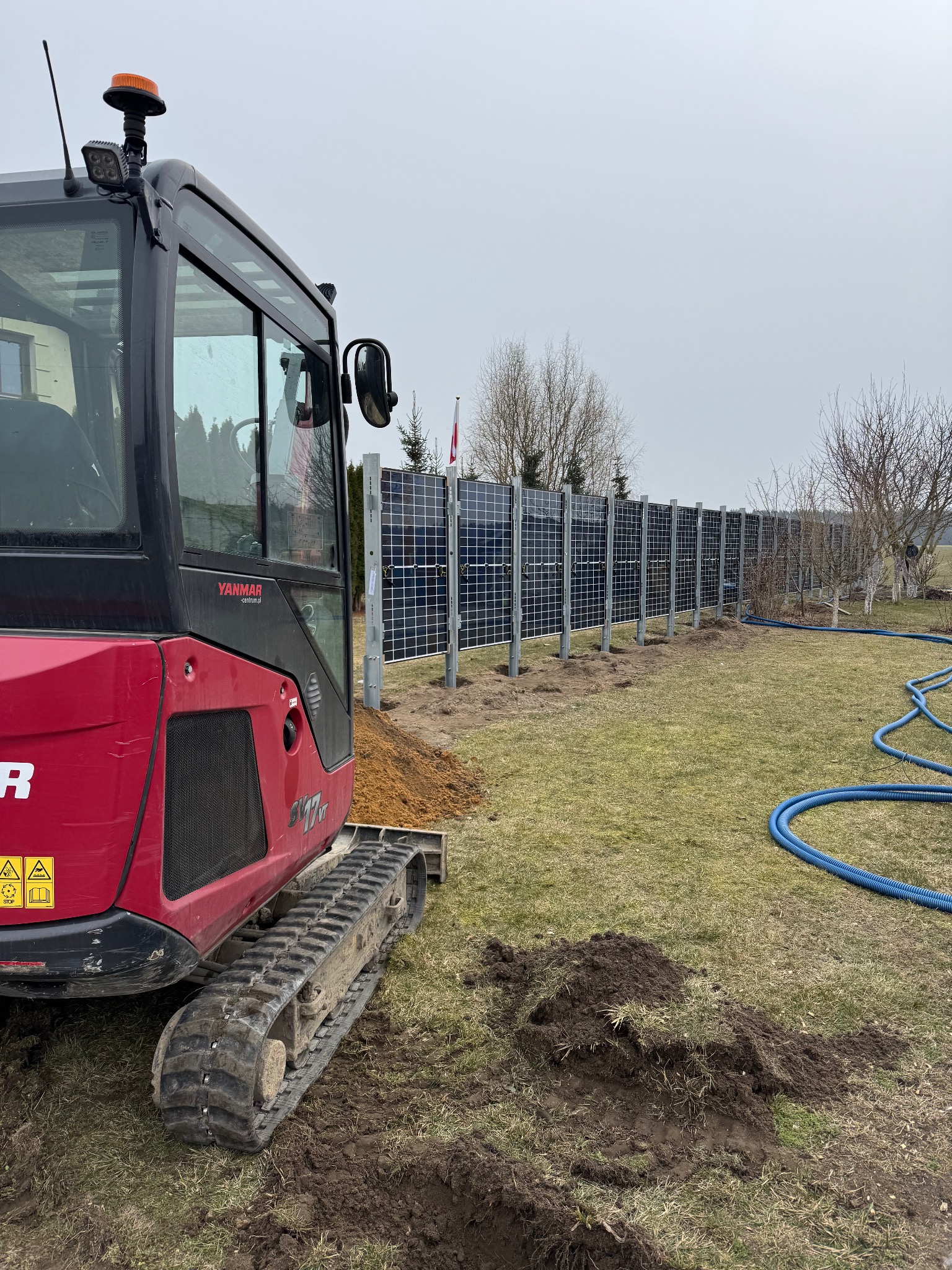 Czerwony minikoparka Yanmar na trawniku obok nowo zainstalowanych paneli fotowoltaicznych, widoczne ślady wykopu i niebieski wąż do instalacji.