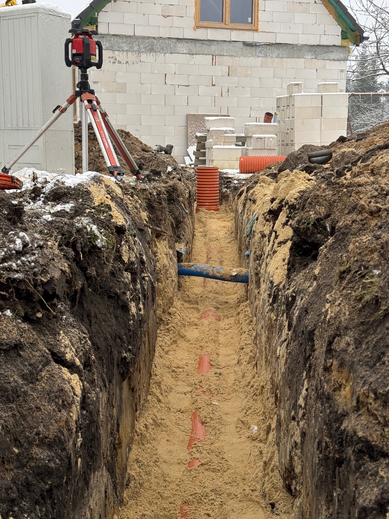 Wąski wykop w piaszczystej ziemi, z widocznymi pomarańczowymi rurami na dnie, częściowo zasypany piaskiem, niebieska rura karbowana na boku, laser krzyżowy na statywie Hilti w tle, budynek...