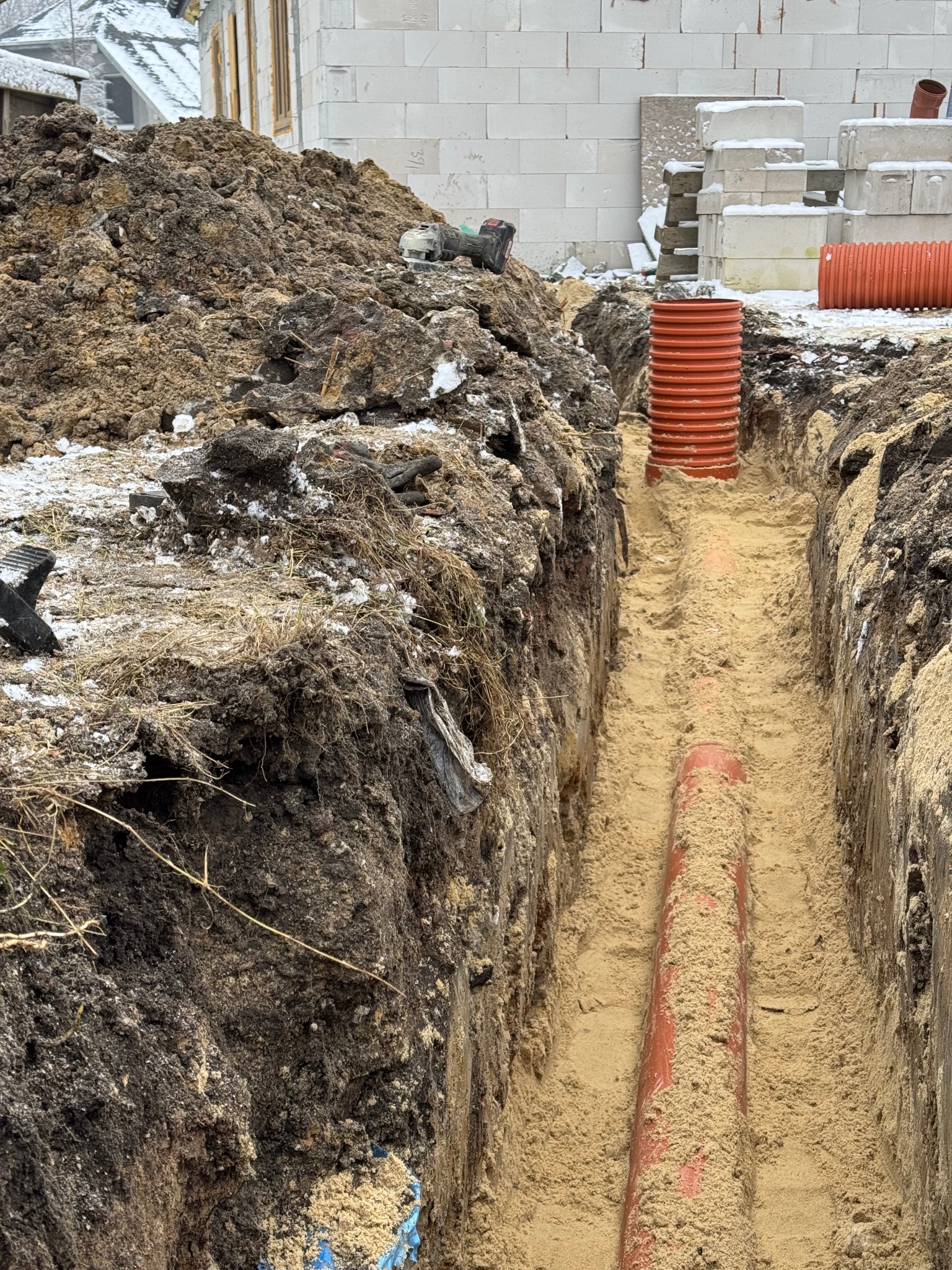 Wykop z ułożoną rurą kanalizacyjną i studzienką rewizyjną z tworzywa sztucznego, obok hałda ziemi z resztkami śniegu i narzędzia budowlane, w tle budynek w stanie surowym.