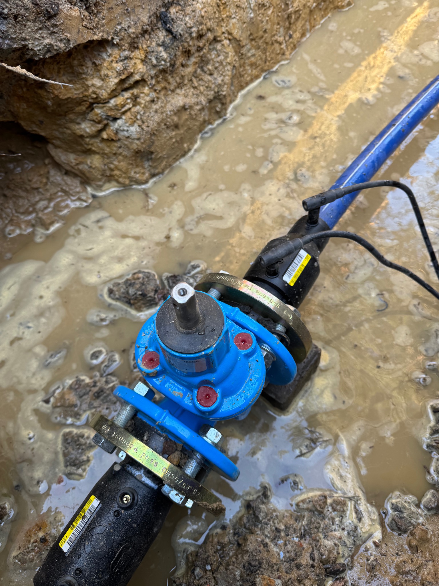 Podłączenie wodociągowe w Bojszowie, widoczne niebieskie rury i zawory w wykopie wypełnionym wodą.