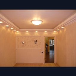 REMBUD - Jasne wnętrze pokoju z oświetleniem punktowym na suficie, dekoracyjną ramą na ścianie z dwoma kinkietami i otwartymi drzwiami do kolejnego pomieszczenia.