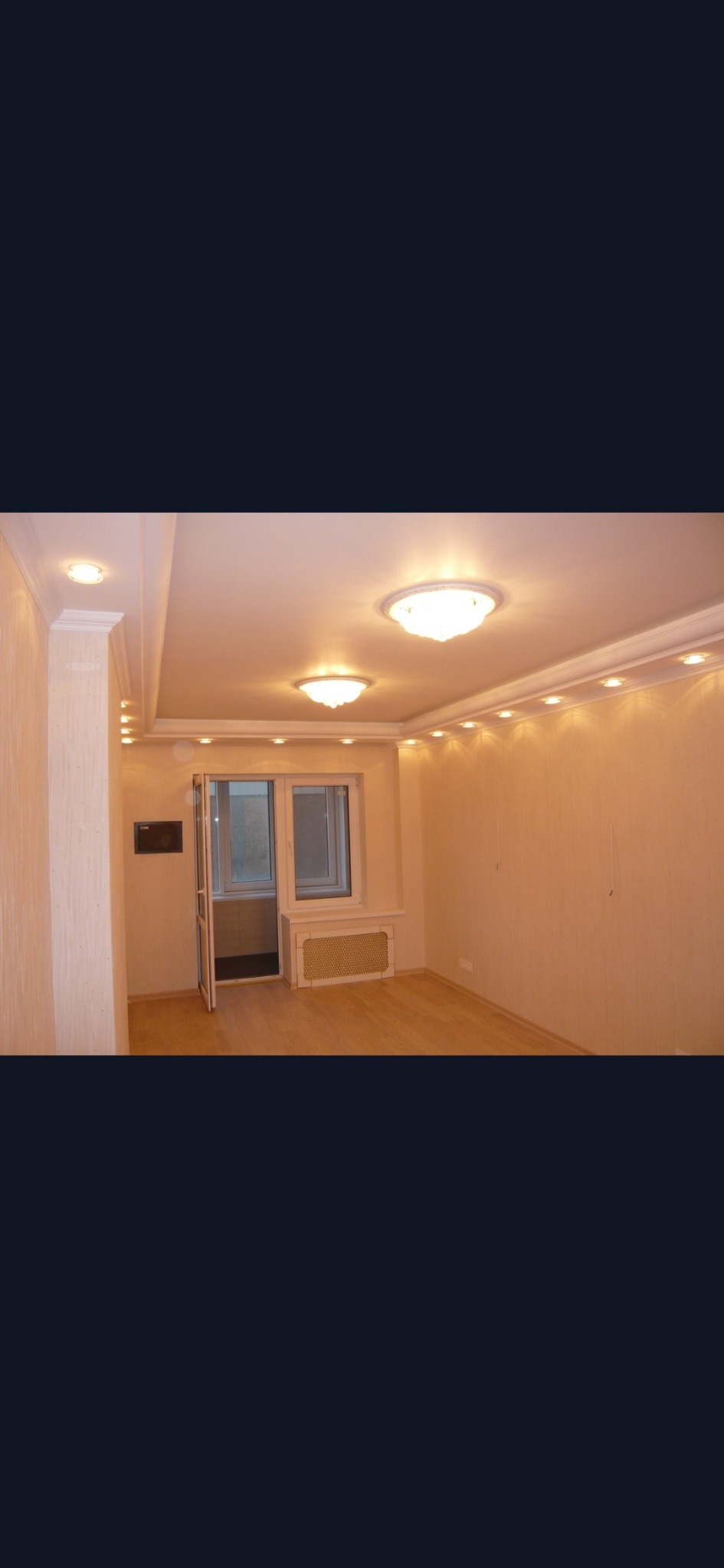 Pusty pokój po remoncie z drewnianą podłogą, kremowymi ścianami, ozdobnym sufitem z oświetleniem punktowym i dwuskrzydłowymi drzwiami balkonowymi.